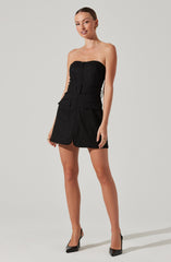 A woman is wearing a strapless black dress with a fitted bodice and a front zipper. The dress features a wide belt with a rectangular buckle around the waist and has large patch pockets on the front of the skirt. She is also wearing black high-heeled shoes.