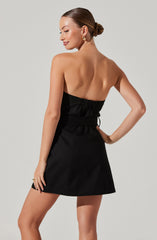 A woman is wearing a strapless black dress with a gathered waistband and a slight A-line skirt. The dress is fitted at the top and flares slightly toward the hem, with the bodice having a ruched design. She has her hair styled in a low bun, and is accessorized with gold hoop earrings and a gold ring.