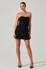 A woman is wearing a strapless black mini dress with a front zipper closure, a wide belt with a rectangular buckle at the waist, and large front flap pockets. She is also wearing black high-heeled shoes.