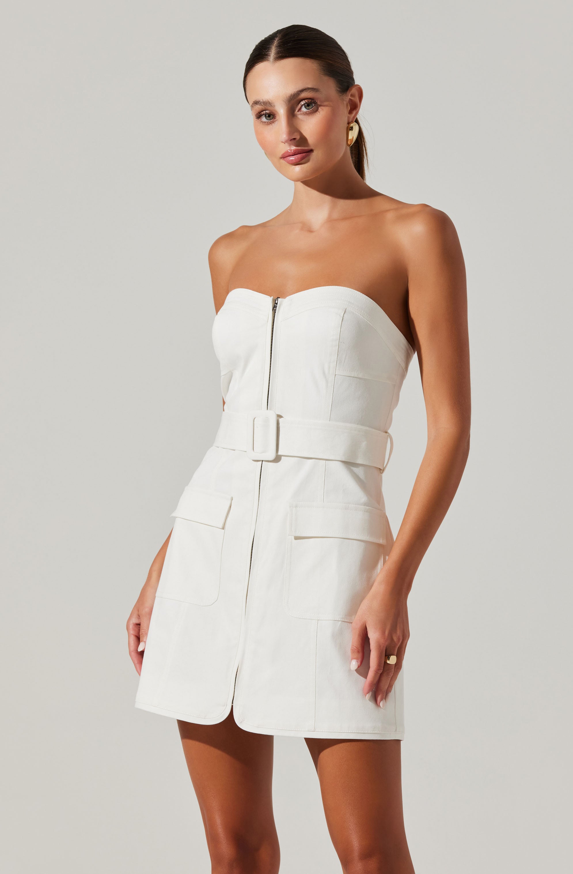 A woman is wearing a strapless, off-white dress with a front zipper and a wide belt at the waist. The dress features large patch pockets on the front. She has her hair pulled back and is accessorized with gold earrings and a ring.