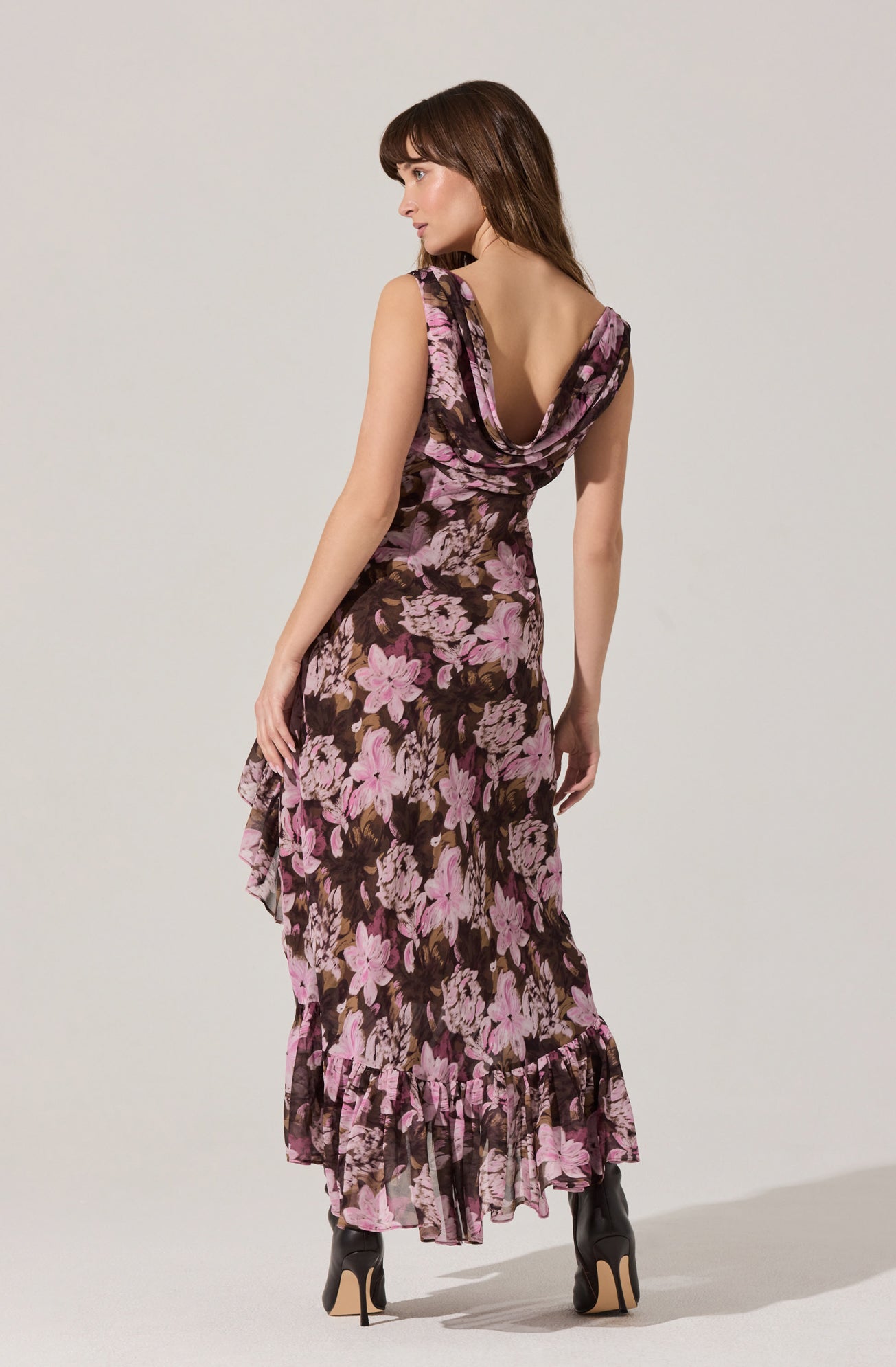 A woman is wearing a sleeveless, floor-length dress with a floral pattern featuring large pink and brown flowers. The dress has a deep cowl neckline and a ruffled hem at the bottom.