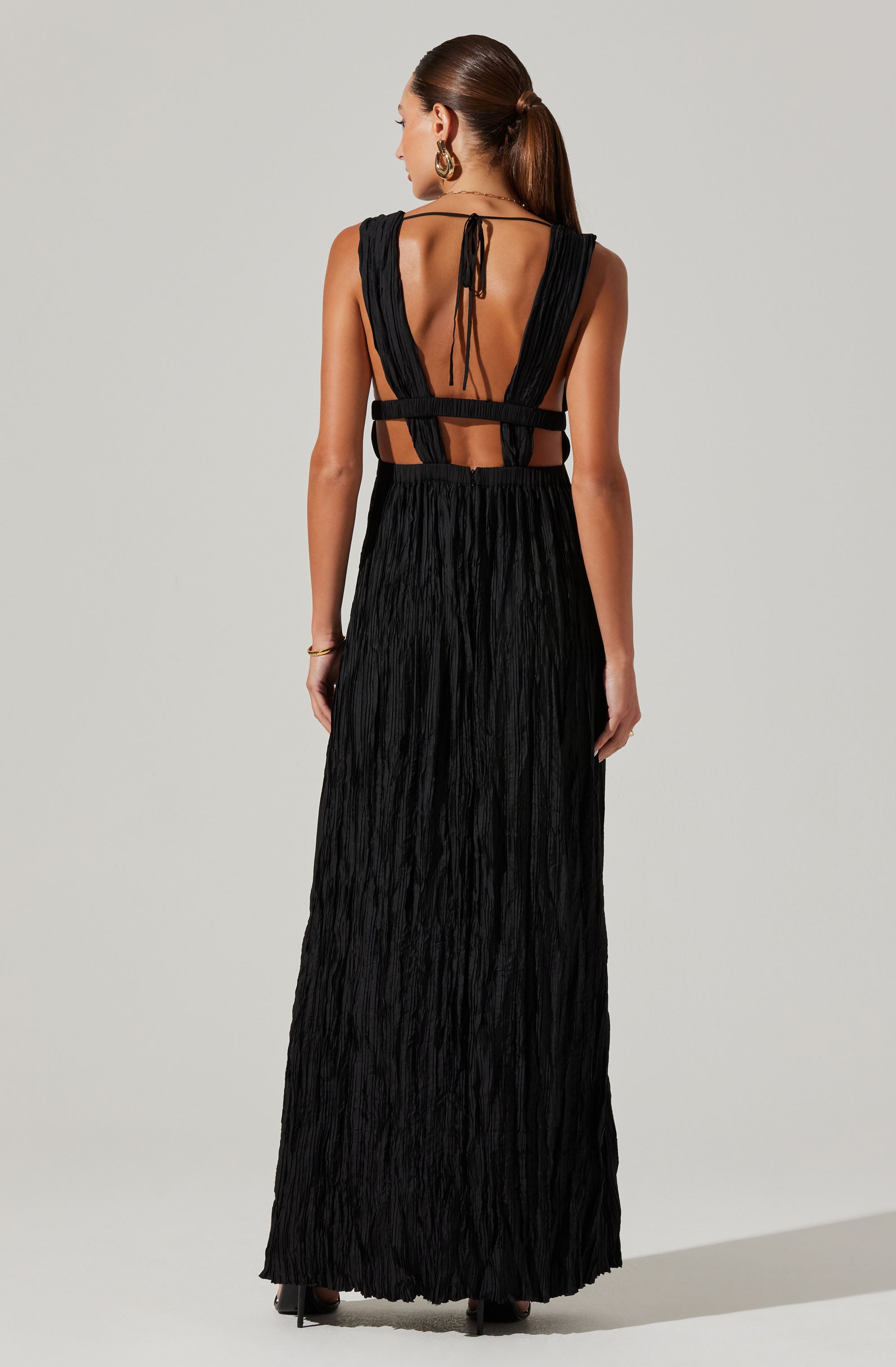 A woman is wearing a long, black dress with a textured, crinkled fabric. The dress features wide shoulder straps that extend over her shoulders and connect at the back with a tie closure. The back of the dress has cut-out sections, with two horizontal straps around the waist and a vertical open back with tied straps at the top. She is accessorized with large hoop earrings and a bracelet on her left wrist.