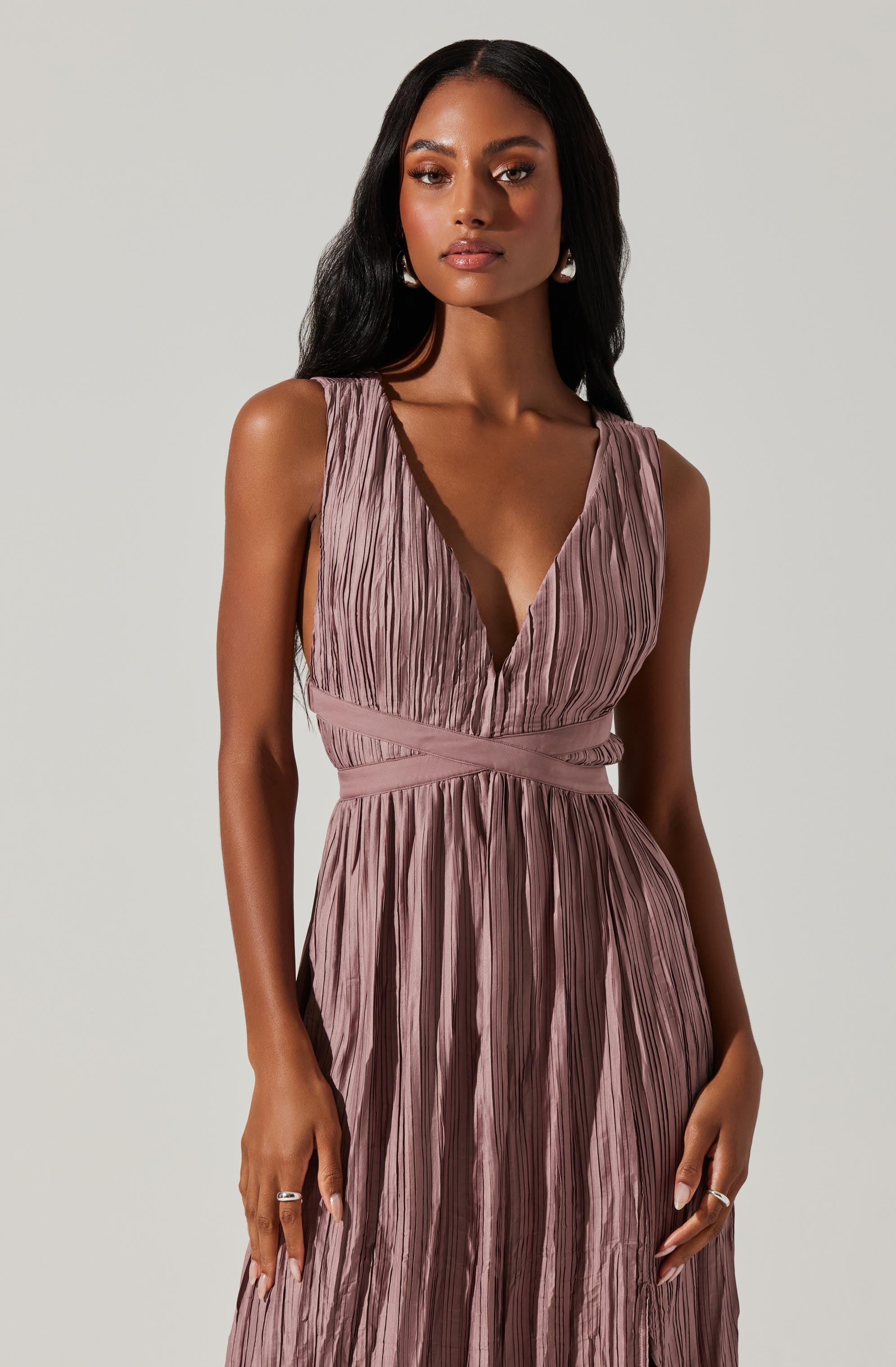 A woman wearing a sleeveless, dusty pink dress with a deep V-neckline. The dress features a crinkled, pleated texture with an empire waist and wide waistband. The skirt falls to the floor with vertical pleats throughout. She accessorizes with dangling pearl earrings and rings on both hands.