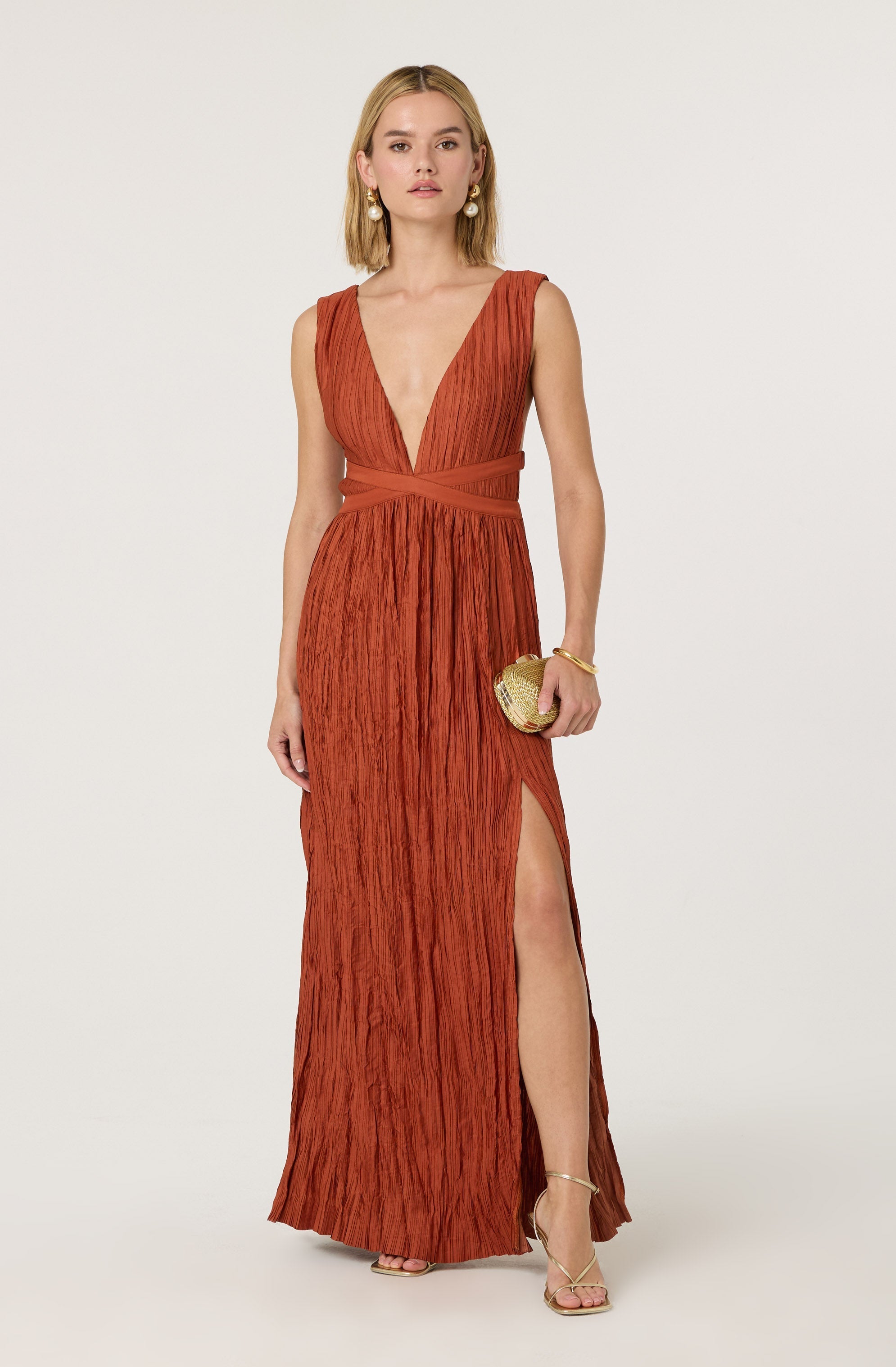 A woman is wearing a floor-length, rust-colored dress with a textured, crinkled fabric. The dress features a deep V-neckline, wide shoulder straps, and a high slit on the left side revealing her leg. She accessorizes with large pearl earrings, a gold bracelet, and carries a small gold clutch. She is also wearing strappy high-heeled sandals.
