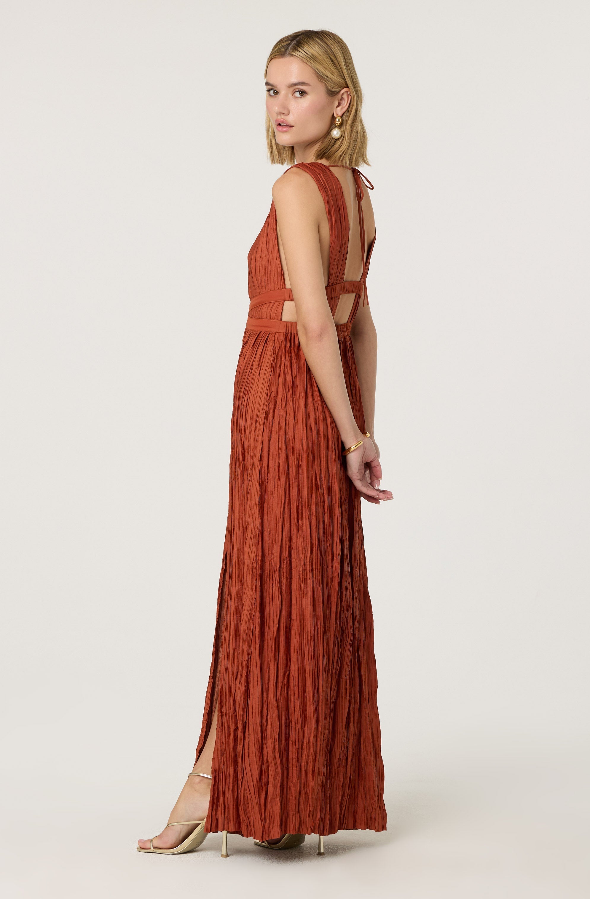 A woman with shoulder-length blonde hair is wearing a long, rust-colored dress with a crinkled texture. The dress features cutout details at the waist and a sheer back with tie closures. She is accessorized with large pearl earrings and a gold bracelet. She is also wearing beige high-heeled sandals.