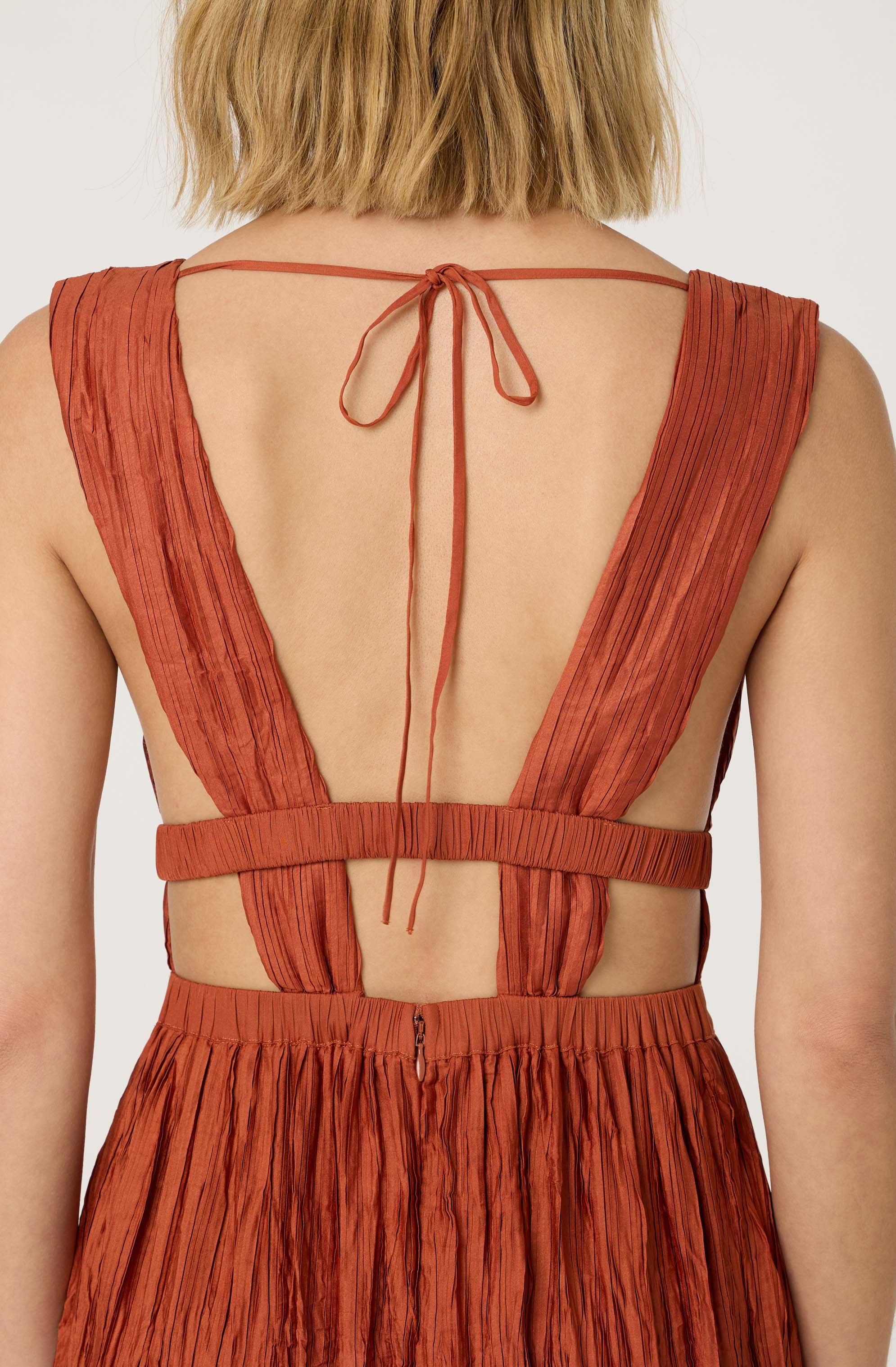 The garment is a sleeveless dress made of a rust-colored, crinkled fabric with vertical pleats. It features wide shoulder straps that extend from the front and tie into a bow at the back. There are cutouts on the sides, connected by a thin horizontal band at the waist and a matching band at the back, which is fastened with a small zipper. The skirt portion of the dress has the same pleated, crinkled texture as the top.