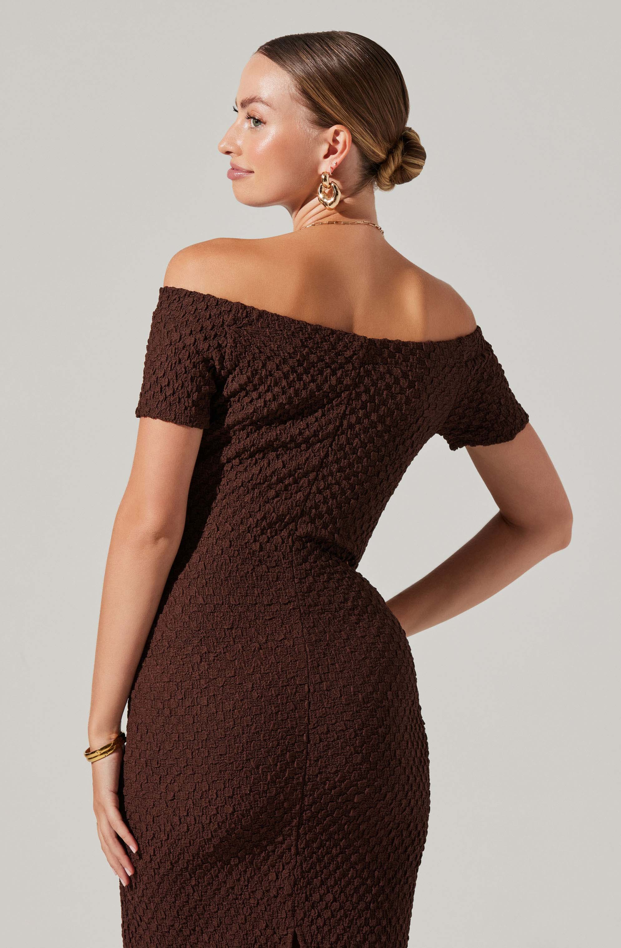 A woman is wearing a brown, textured, off-the-shoulder dress with short sleeves. The dress has a fitted silhouette, and she accessorizes with large, gold hoop earrings, a delicate chain necklace, and a bracelet. Her hair is styled in a sleek bun.