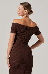 A woman is wearing a brown, textured, off-the-shoulder dress with short sleeves. The dress has a fitted silhouette, and she accessorizes with large, gold hoop earrings, a delicate chain necklace, and a bracelet. Her hair is styled in a sleek bun.