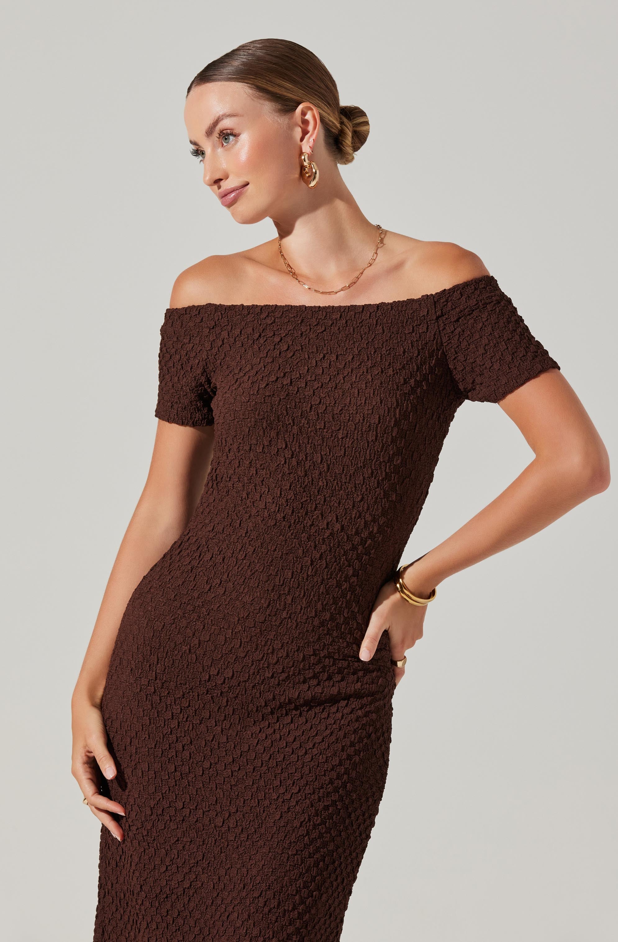 A woman is wearing an off-the-shoulder dress with a textured, waffle-knit pattern in a rich chocolate brown color. The dress is fitted and falls to below the knee. She accessorizes with gold hoop earrings, a delicate gold necklace, and gold bracelets.