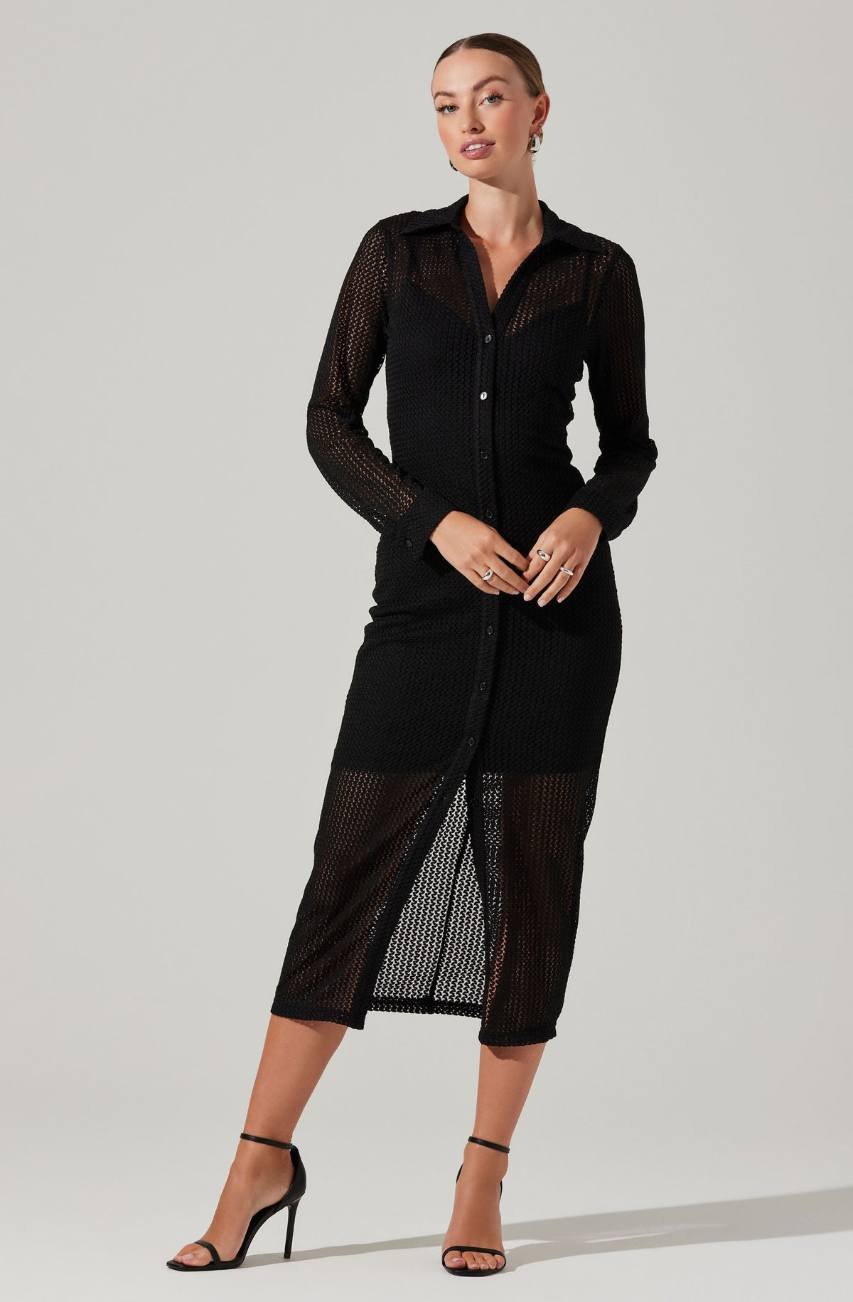 A woman is wearing a black, long-sleeved button-up dress made of a see-through, mesh fabric with a striped pattern. The dress has a collar and a front slit that reveals her legs. She is also wearing black high-heeled sandals with ankle straps.