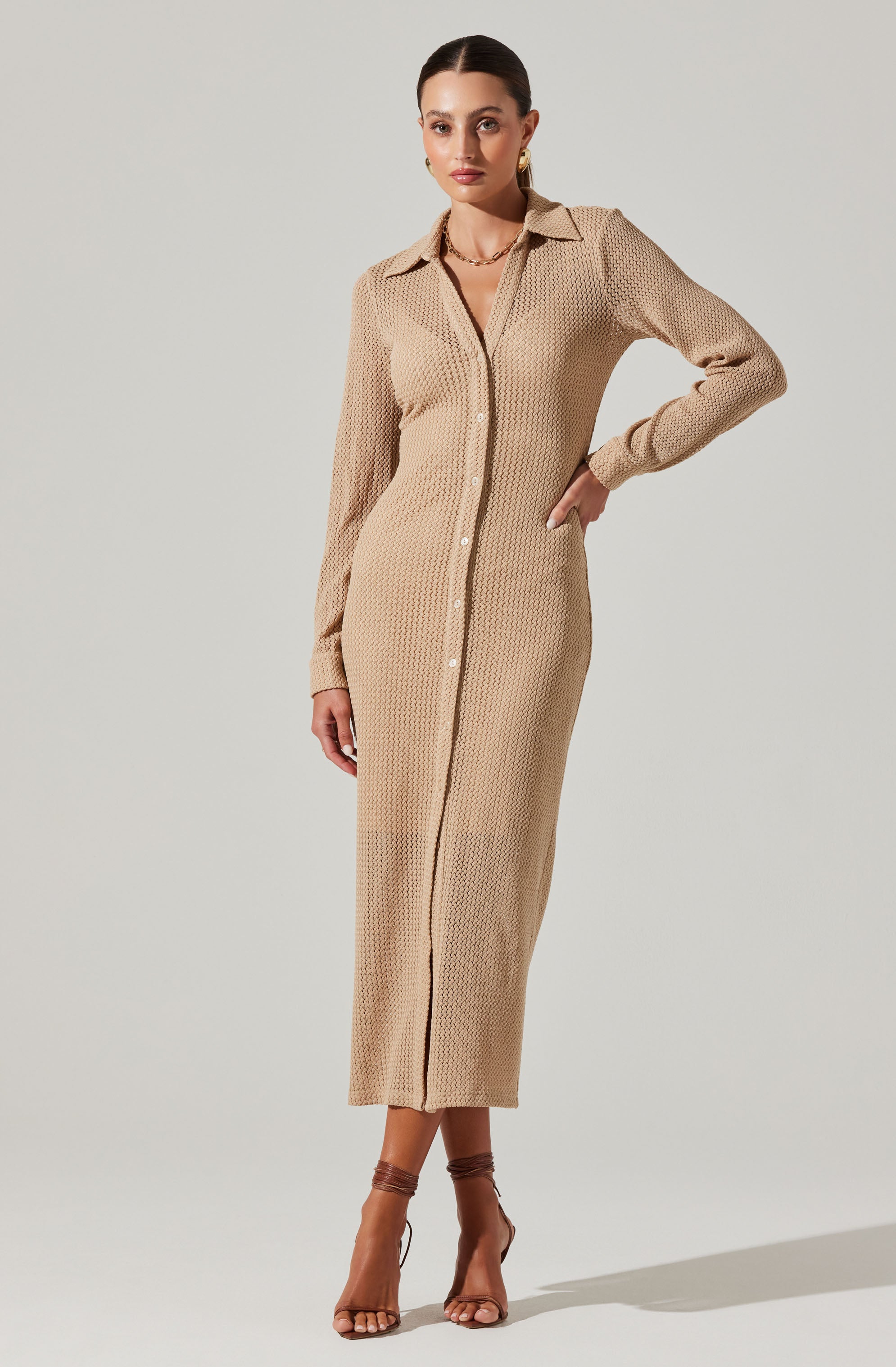 A woman is wearing a long beige shirt dress made of a textured, knit fabric. The dress features a classic collar, a button-down front with white buttons, and long sleeves with cuffs. She is also wearing strappy high-heeled sandals.