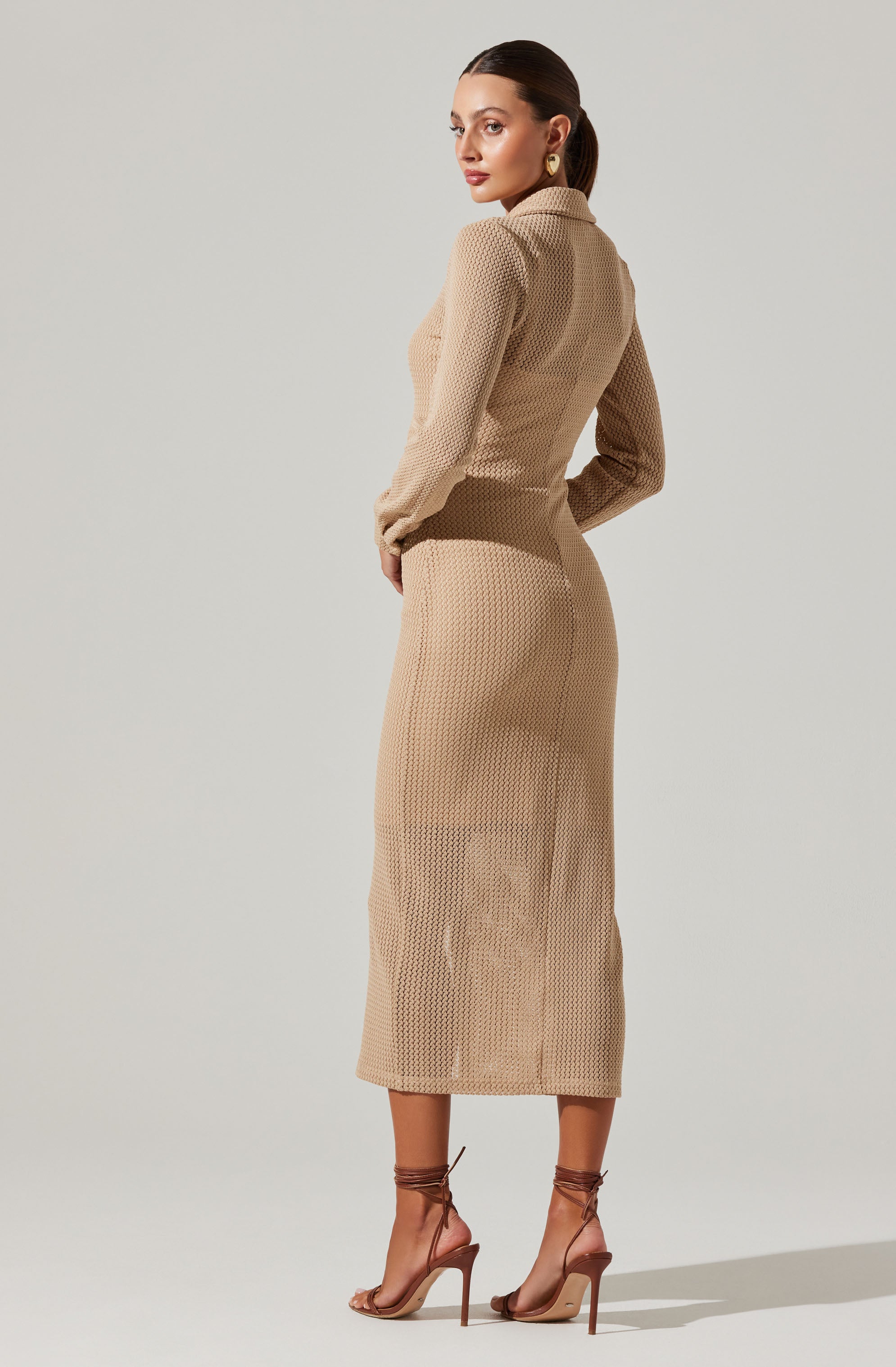 A woman is wearing a beige, textured knit long-sleeve top with a collar, paired with a matching high-waisted, knit midi skirt. She has on nude strappy high-heeled sandals that lace up around her ankles and gold hoop earrings.