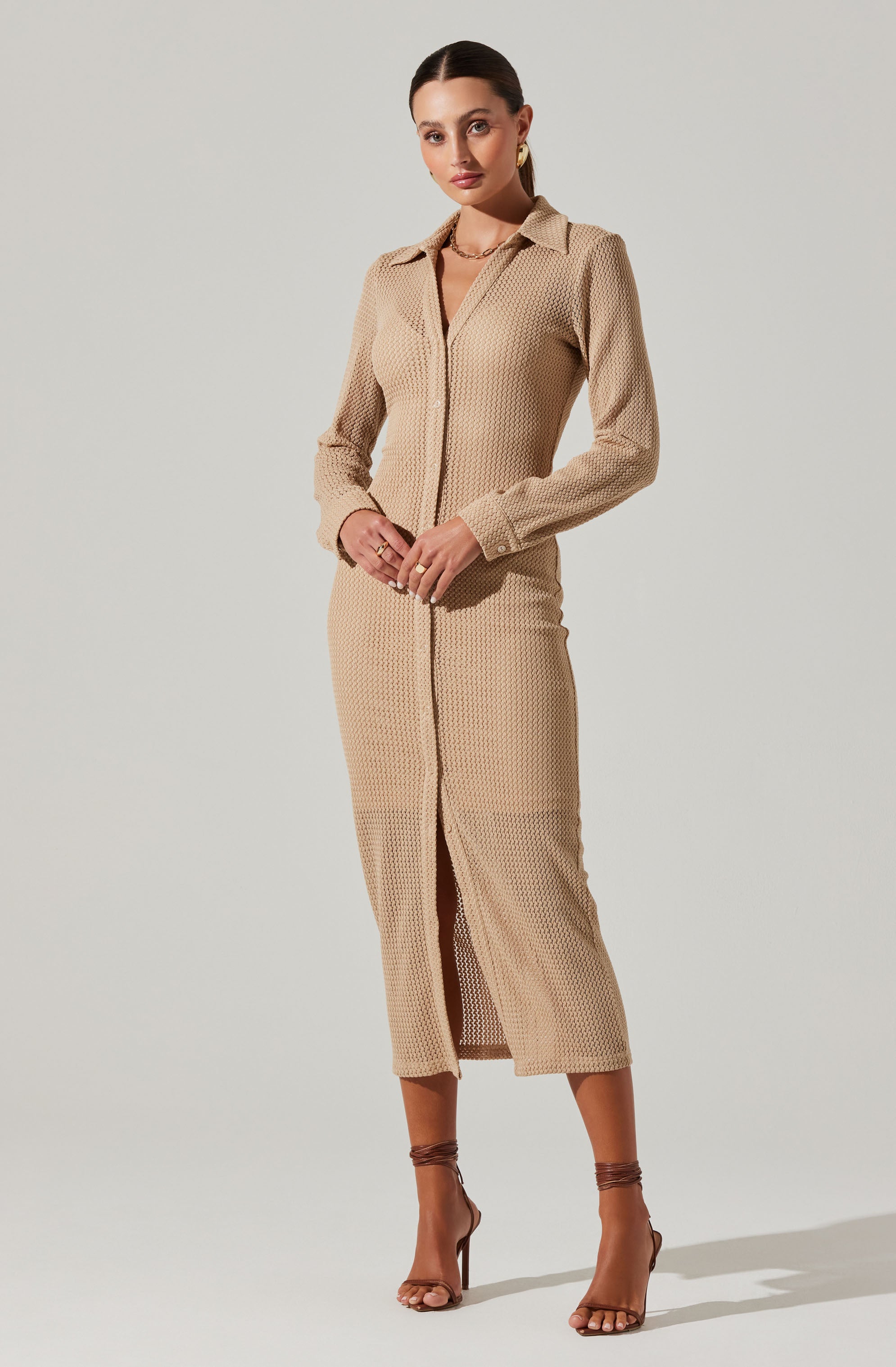 A woman is wearing a long beige knit dress with a subtle textured pattern. The dress features a collar and a front zipper closure that runs from the neckline to the hem, with a small slit at the bottom front. She is accessorized with gold hoop earrings and layered necklaces, and is wearing strappy high-heeled sandals.