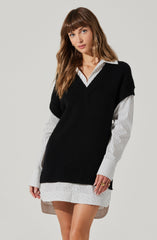 A woman is wearing a white button-up shirt with thin black vertical stripes, layered with an oversized black knit vest. The shirt's collar and cuffs are visible, and the shirt's hem extends below the vest, creating a layered look. The woman has long, wavy brown hair and is standing against a plain light-colored background.