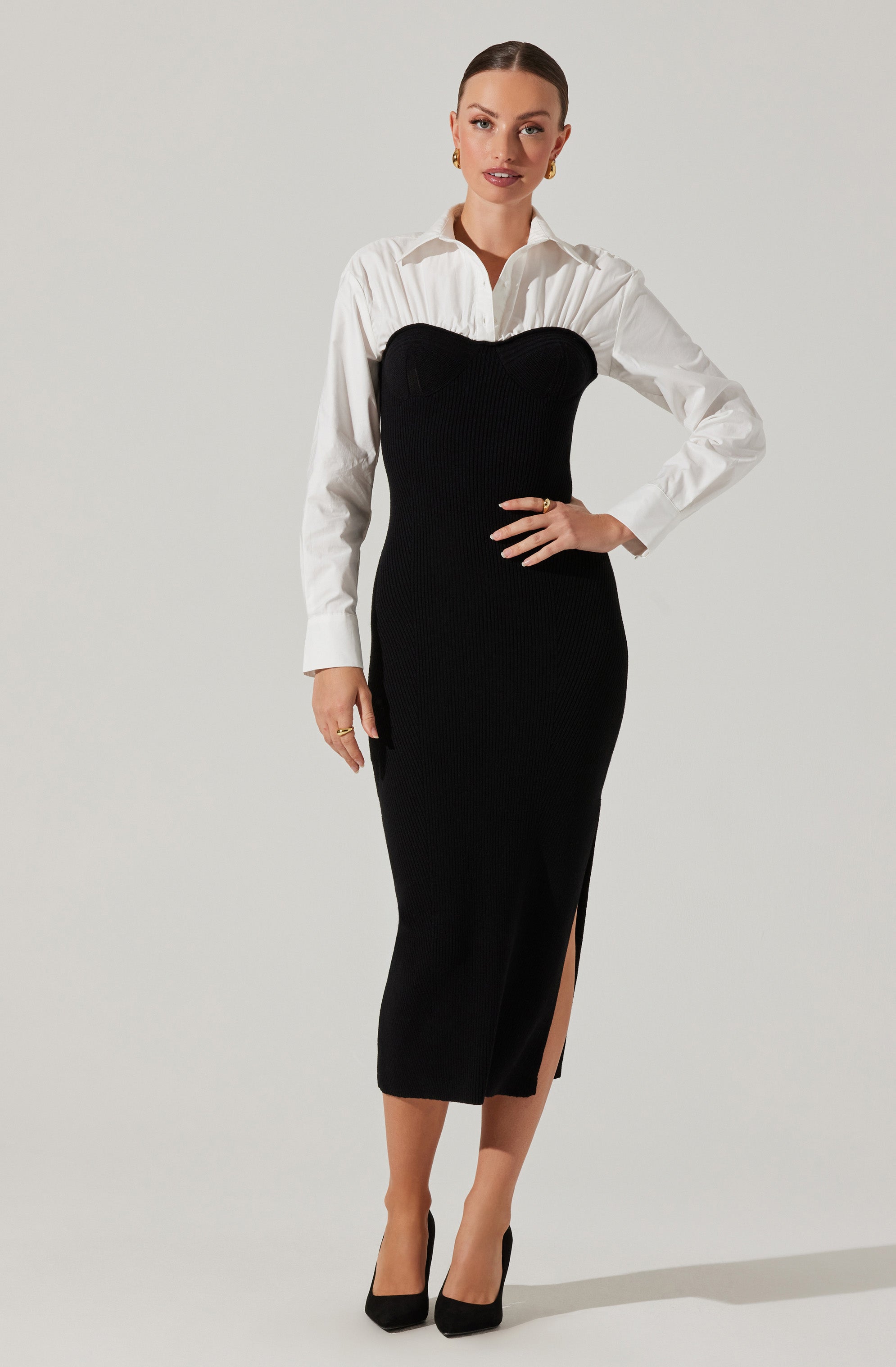 A woman is wearing a white long-sleeved button-up shirt underneath a black, strapless, ribbed knit dress that extends to mid-calf. The dress features a high slit on one side. She is also wearing black high-heeled shoes and gold jewelry, including earrings and rings.