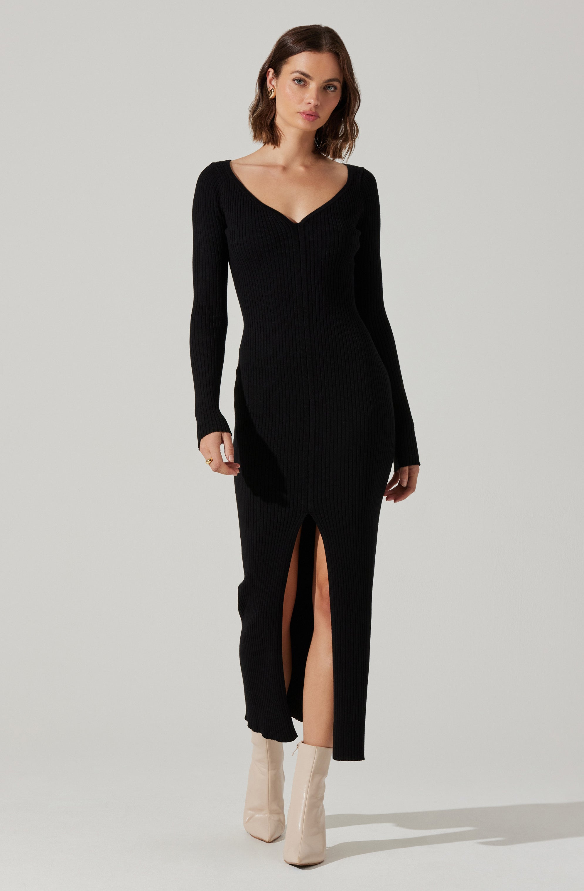 A woman is wearing a long, fitted black ribbed knit dress with a V-neckline and long sleeves. The dress features a front slit that extends below the knee, revealing her legs and beige ankle boots.