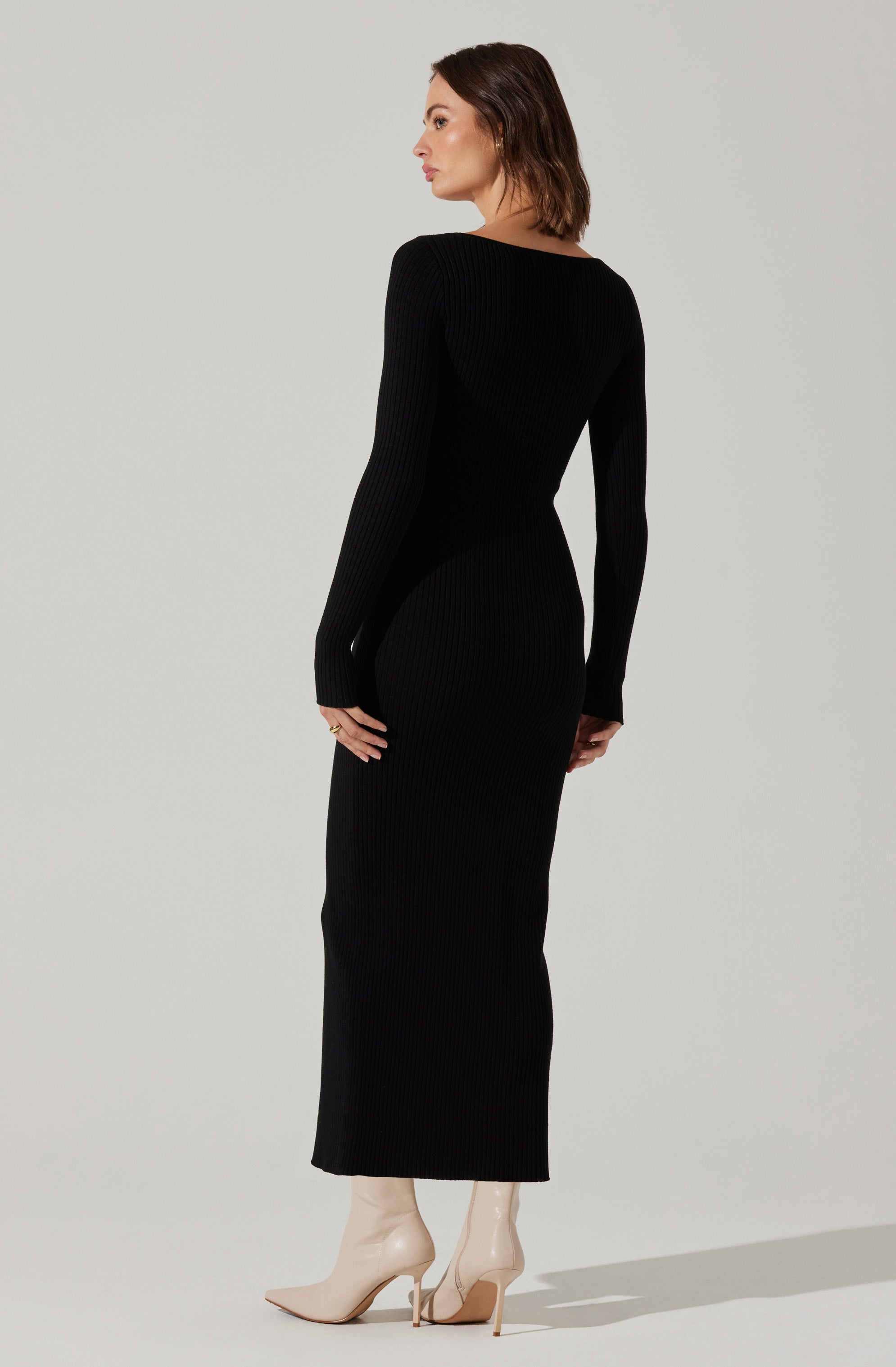 A woman is wearing a long, form-fitting, black ribbed knit dress with long sleeves and a high neckline. She pairs the outfit with beige high-heeled boots.