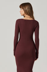 A woman is wearing a form-fitting, long-sleeved ribbed dress in a deep burgundy color. The dress has a crew neckline and features vertical ribbed knitting throughout. She is accessorized with a simple gold chain necklace.