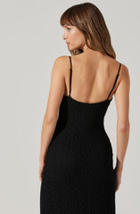 A woman is wearing a black, textured, sleeveless dress with thin adjustable straps. The dress fits closely to her body and has a scoop back.