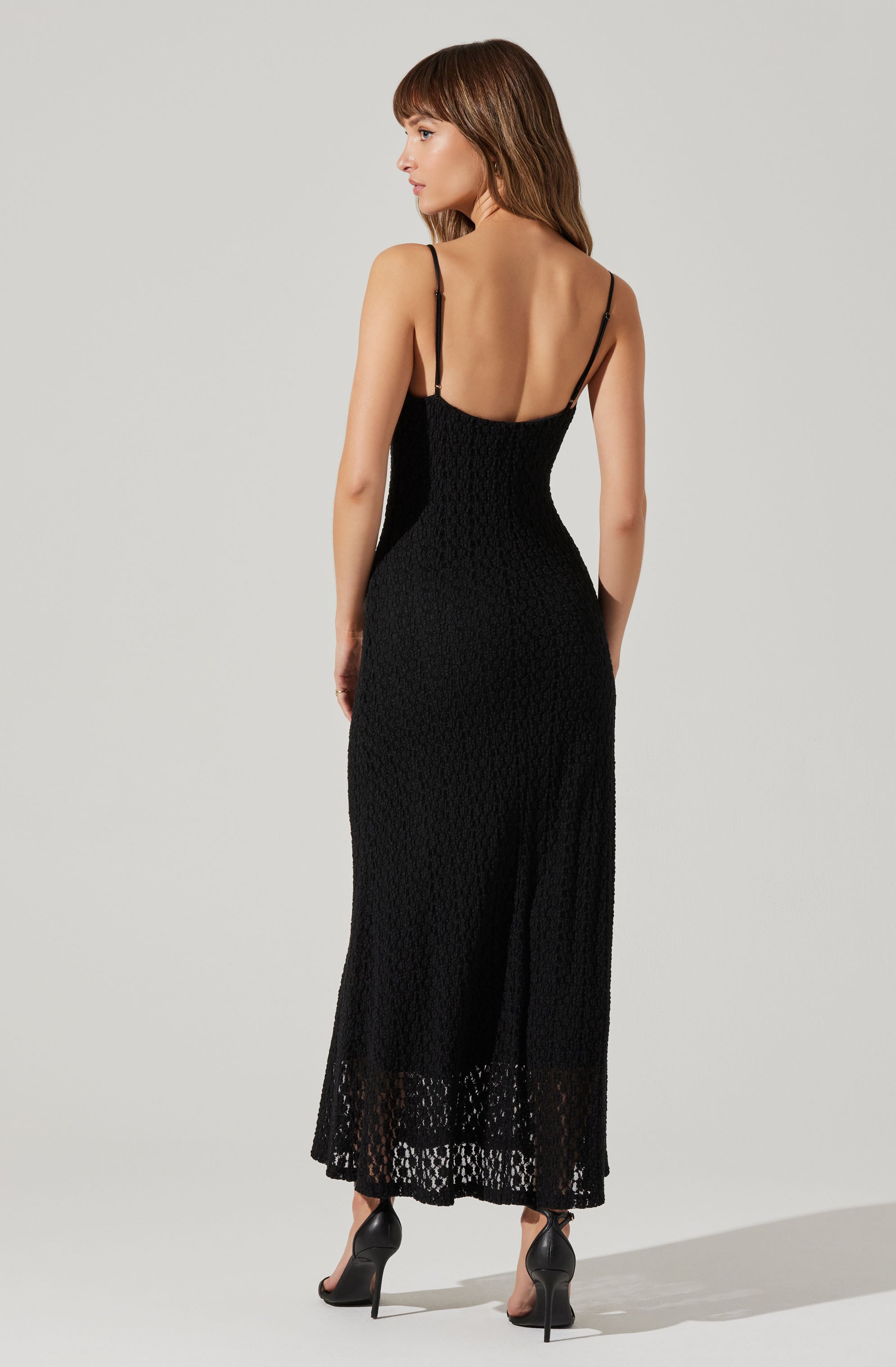 A woman is wearing a long black dress with thin spaghetti straps, fitted at the waist and hips, and featuring a textured, lace-like pattern throughout. The dress reaches down to her ankles with a slight flare at the hem. She is also wearing black high-heeled shoes.