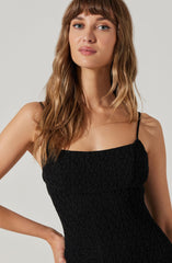 A woman is wearing a fitted black dress with a textured, smocked fabric. The dress has thin spaghetti straps and a square neckline.