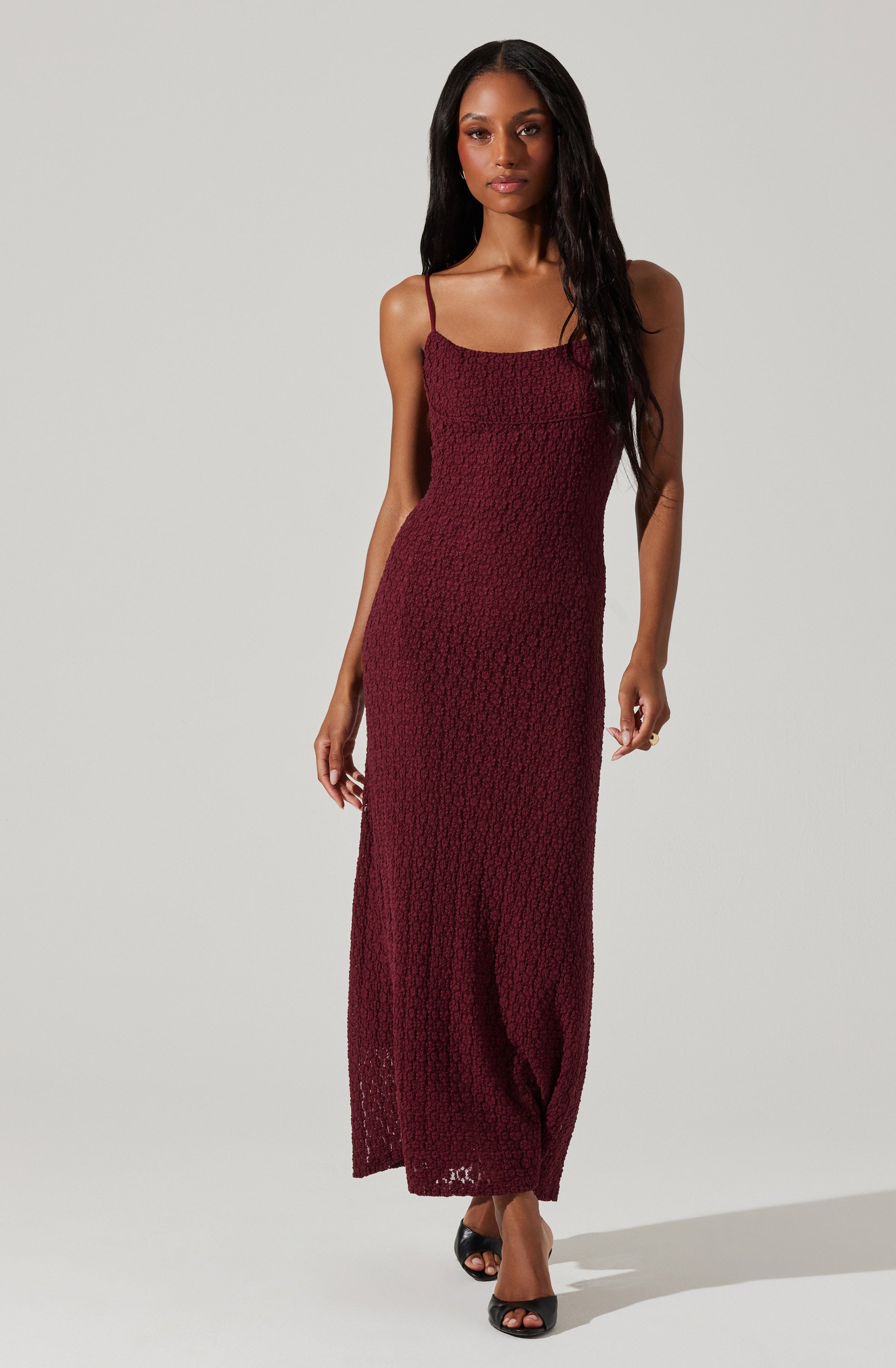 A woman is wearing a long, sleeveless, burgundy dress with thin spaghetti straps. The dress features a textured, smocked fabric that extends from the bust to the hem, creating a fitted silhouette. She is pairing the dress with open-toed black heels.