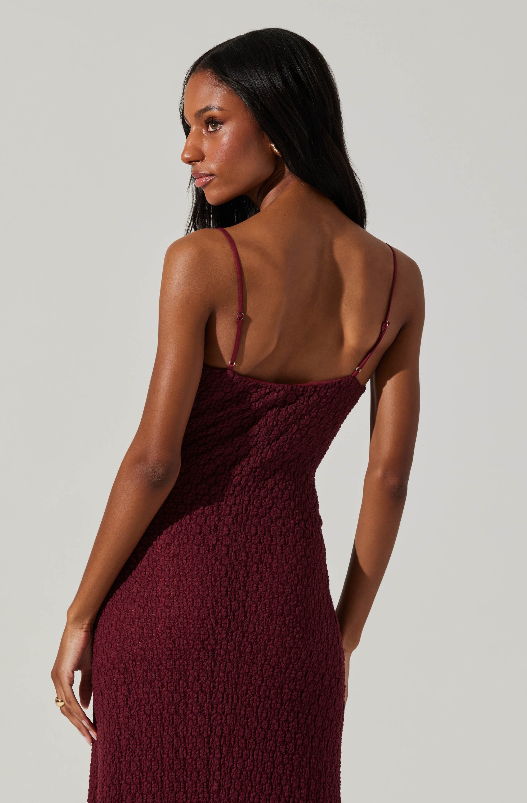 A woman is wearing a sleeveless, burgundy-colored dress with thin spaghetti straps. The dress has a textured, smocked fabric and fits snugly, highlighting her figure.