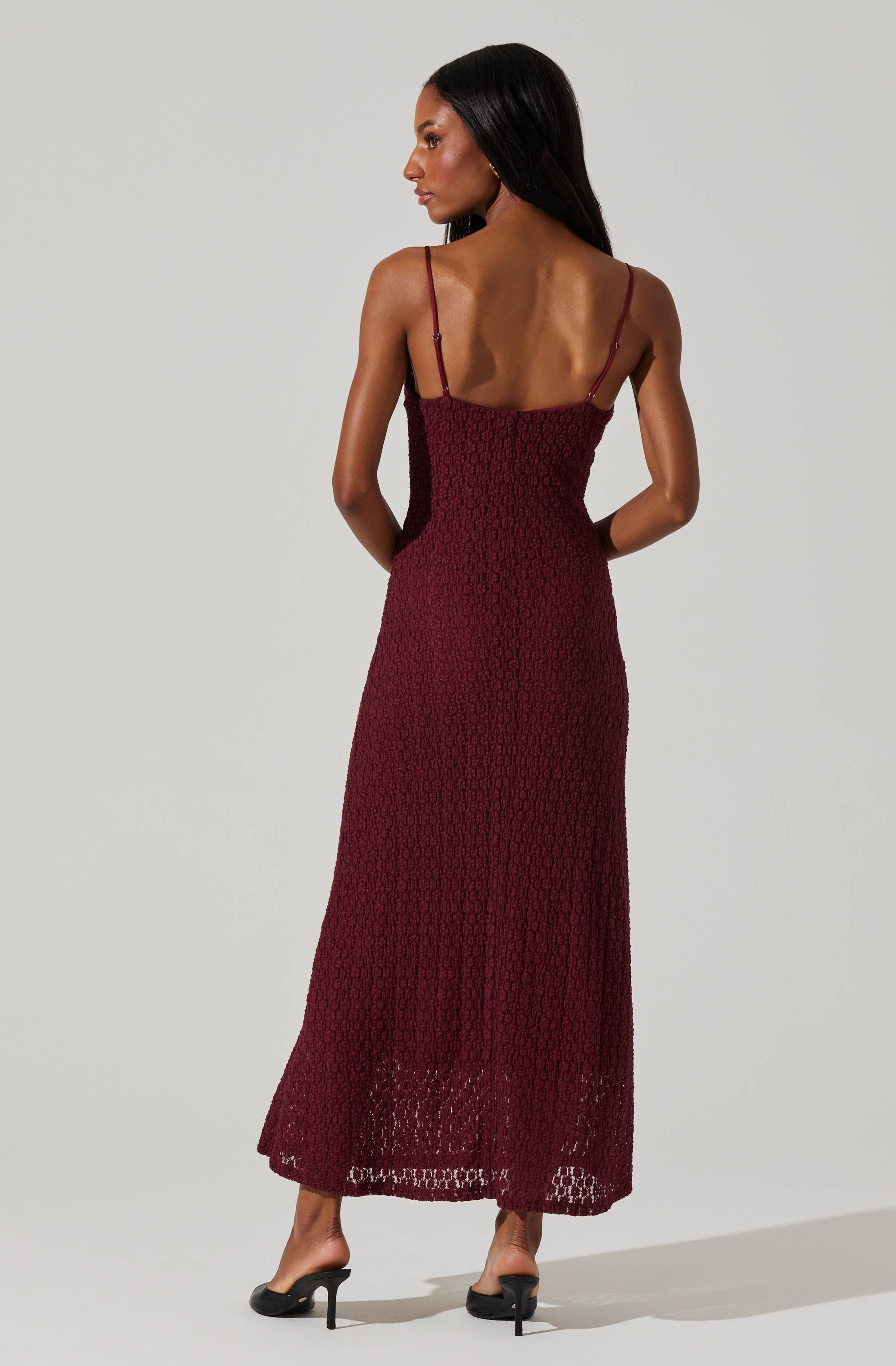 A woman is wearing a long, sleeveless burgundy dress with thin shoulder straps. The dress has a textured, crochet-like pattern throughout and reaches down to her ankles. She is also wearing black high-heeled sandals.