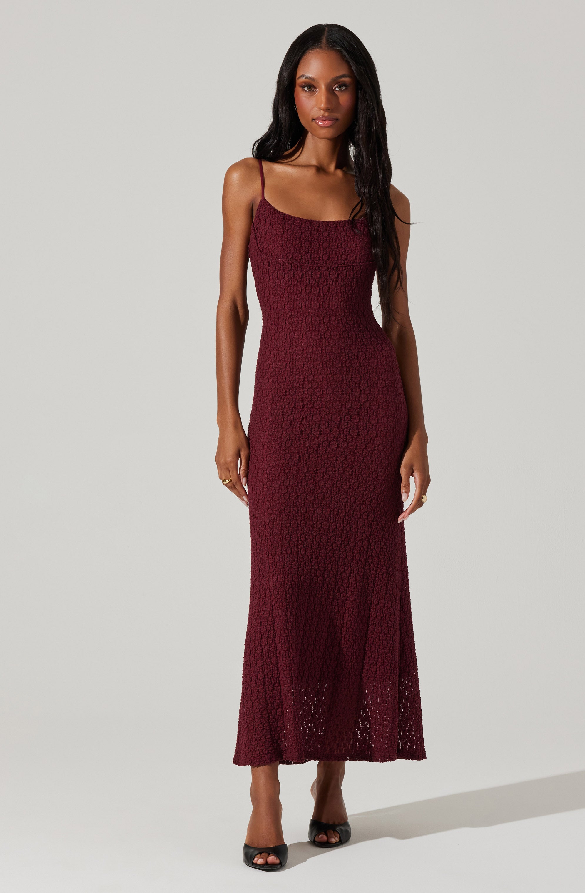 A woman is wearing a sleeveless, full-length dress with spaghetti straps. The dress is made of a textured, crochet-like fabric in a deep burgundy color. She is also wearing open-toe black heels.