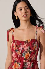 A woman is wearing a sleeveless dress with shoulder straps tied into bows on each shoulder. The dress features a colorful, abstract print in shades of red, orange, pink, green, and white, with a gathered, ruched bodice and a ruffled waistband. She has long, dark hair and is accessorized with large, gold, shell-shaped earrings.