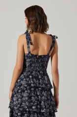 A woman is wearing a sleeveless dress with shoulder straps tied into bows. The dress features a dark base color with a lighter floral pattern and has a layered, tiered design.