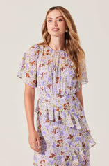 A woman is wearing a matching floral outfit made of lightweight, semi-sheer fabric. The top features short, puffed sleeves and a round neckline, with a loose fit. The skirt has a tiered, ruffled design with a flowing silhouette, and the pattern consists of abstract floral prints in shades of lavender, purple, reddish-brown, and white.