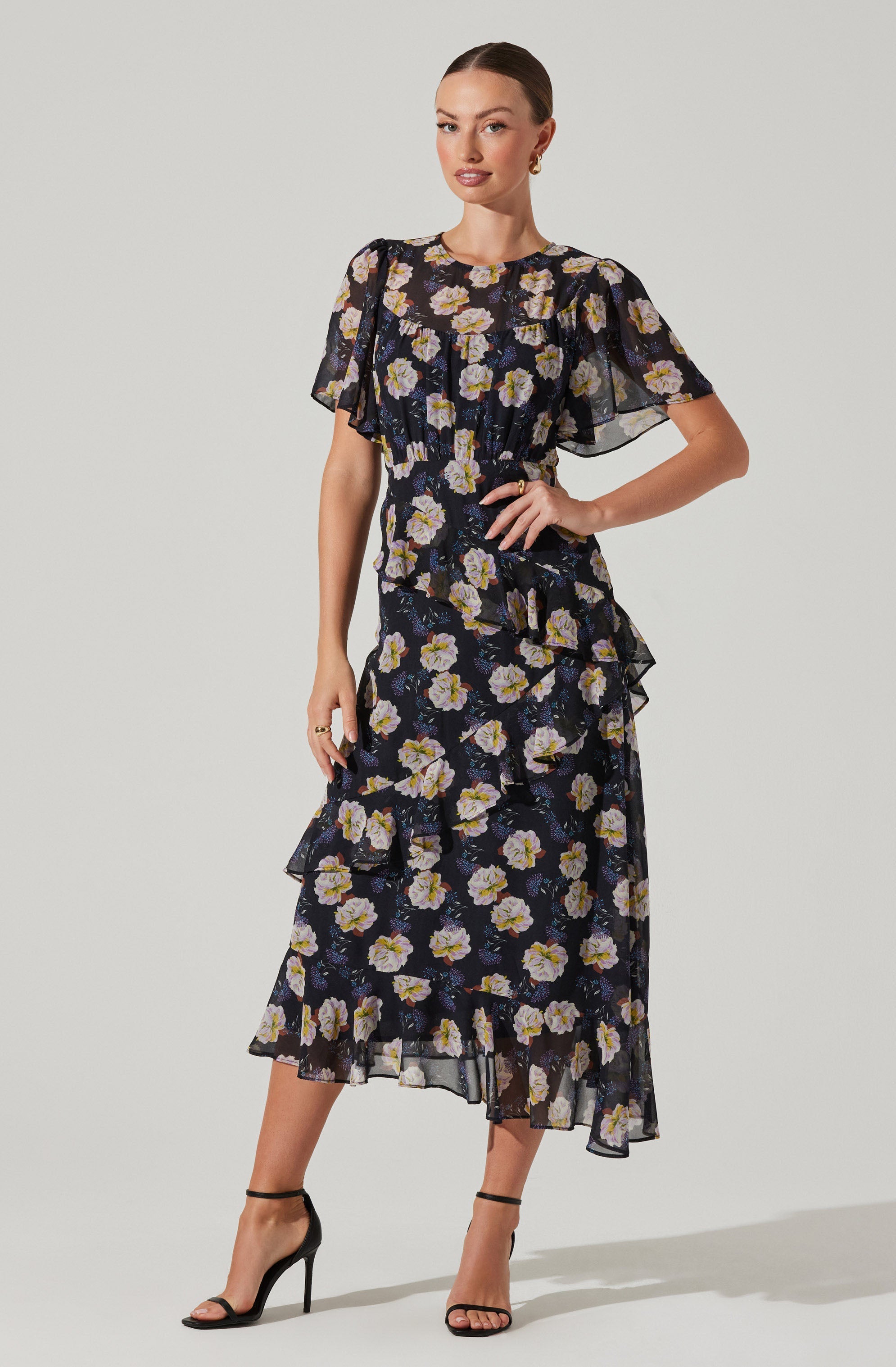 A woman is wearing a two-piece dress set with a floral pattern. The top is a short-sleeved, black sheer fabric with large, light-colored flowers and ruffle details on the sleeves and around the waist. The skirt is a matching black sheer fabric with the same large floral pattern, featuring ruffles along the layers and an asymmetrical hemline. She is also wearing black high-heeled sandals with ankle straps.