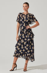 A woman is wearing a two-piece dress set with a floral pattern. The top is a short-sleeved, black sheer fabric with large, light-colored flowers and ruffle details on the sleeves and around the waist. The skirt is a matching black sheer fabric with the same large floral pattern, featuring ruffles along the layers and an asymmetrical hemline. She is also wearing black high-heeled sandals with ankle straps.