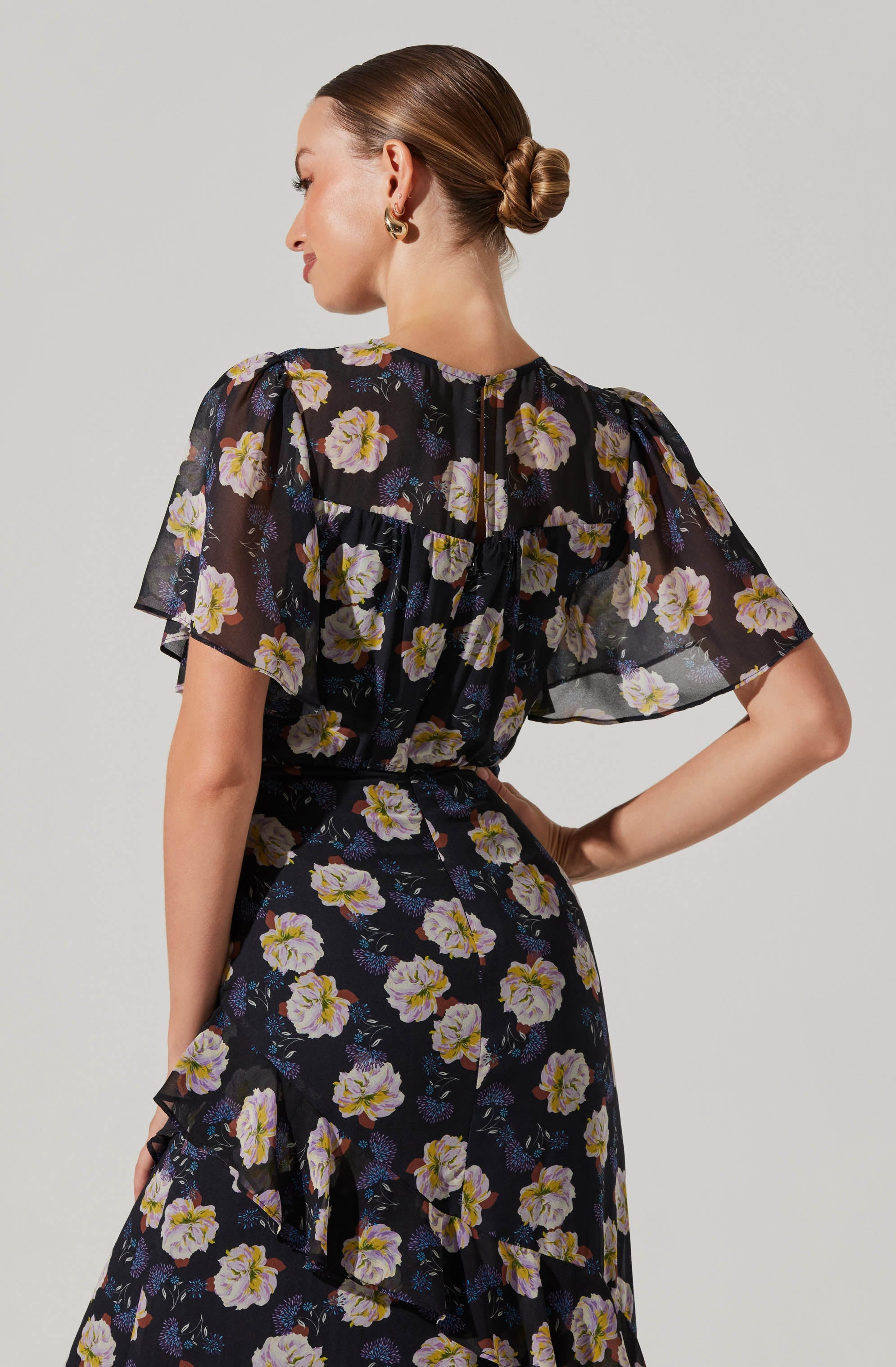 A woman wearing a black dress with a floral pattern consisting of white and yellow flowers with purple accents. The dress has short, sheer, ruffled sleeves and a cinched waist.