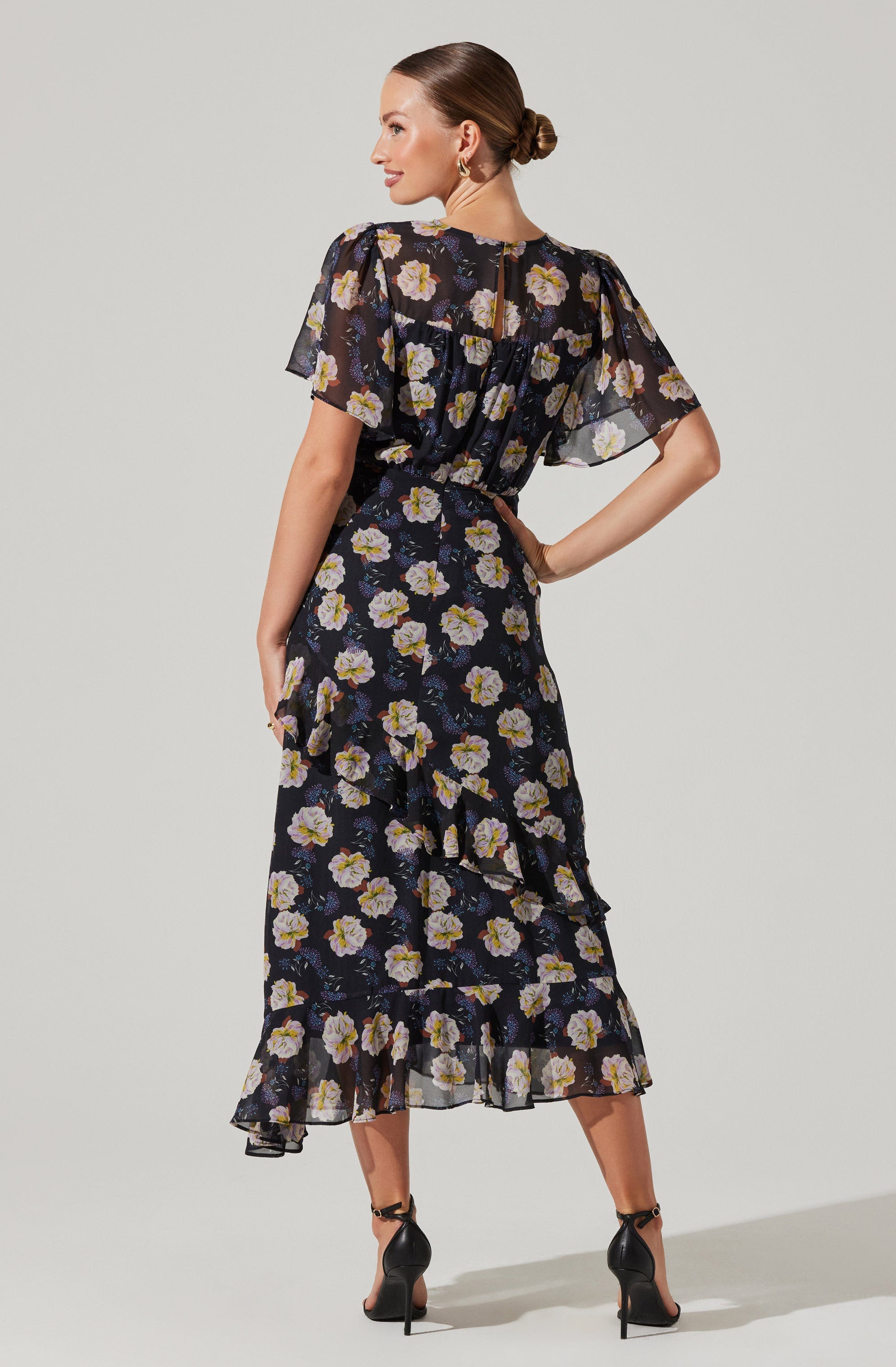 A woman is wearing a black dress with a floral print featuring light purple, yellow, and white flowers. The dress has short, sheer, puffed sleeves and a high waist. It has a tiered, ruffled skirt that reaches mid-calf length. The dress appears lightweight and flowy. She is also wearing black high-heeled sandals with ankle straps.