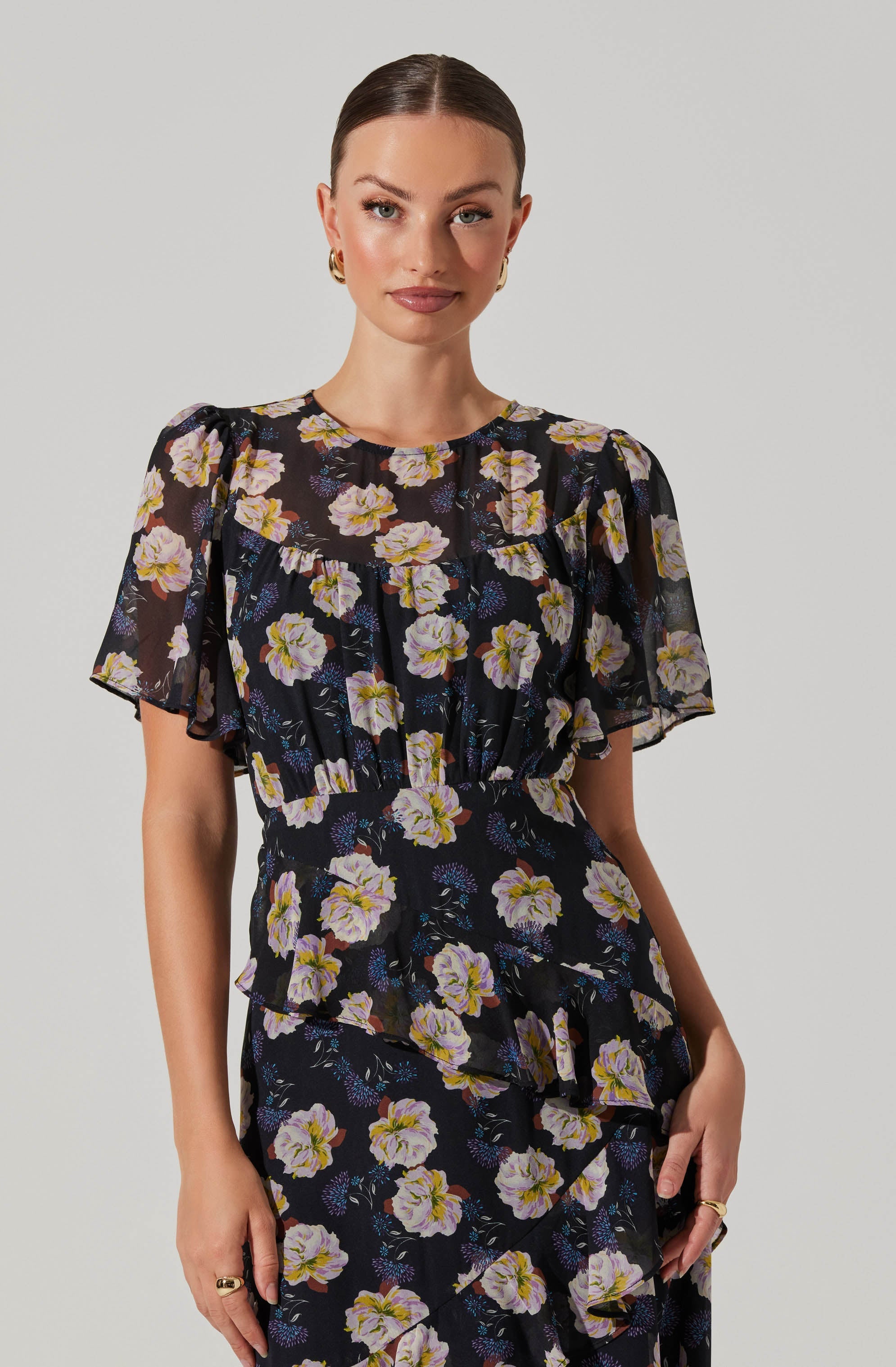 A woman is wearing a black dress featuring a floral pattern with white, lavender, and yellow flowers. The dress has short, sheer sleeves with a slightly puffed design, a round neckline, and a fitted waist. The fabric appears lightweight and flowy, with a ruffled detail running diagonally across the skirt.