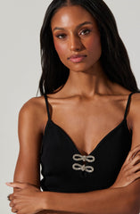 A woman with long dark hair is wearing a black sleeveless top with thin spaghetti straps. The top features a decorative bow detail made of sparkling material at the center of the neckline.