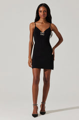 A woman is wearing a black sleeveless dress with thin spaghetti straps. The dress has a fitted silhouette and features a decorative metallic clasp detail at the front center. She is also wearing black high-heeled shoes with ankle straps.