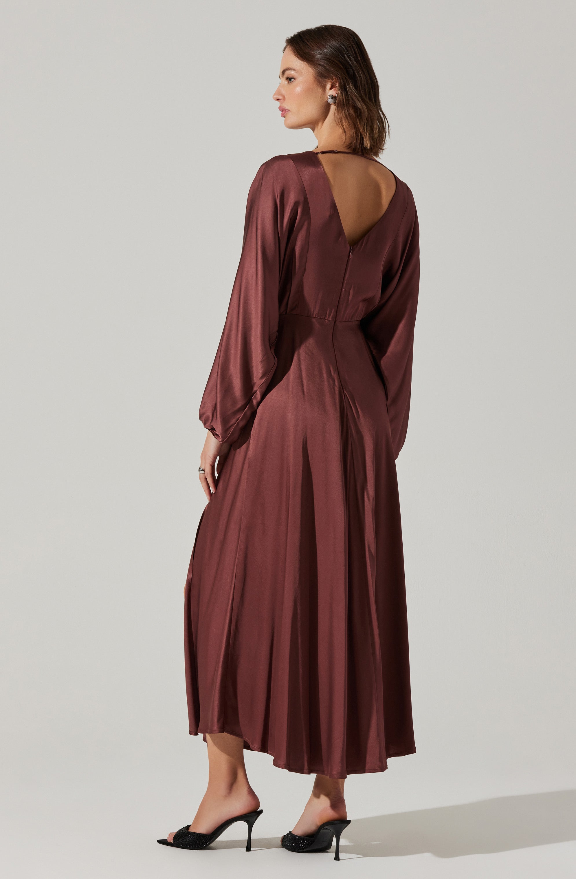 A woman is wearing a long, satin, burgundy dress with long billowy sleeves that gather at the wrists. The dress has a deep V-shaped back neckline and a fitted waist, flowing into a pleated, ankle-length skirt. She is paired with black high-heeled sandals that have thin straps.