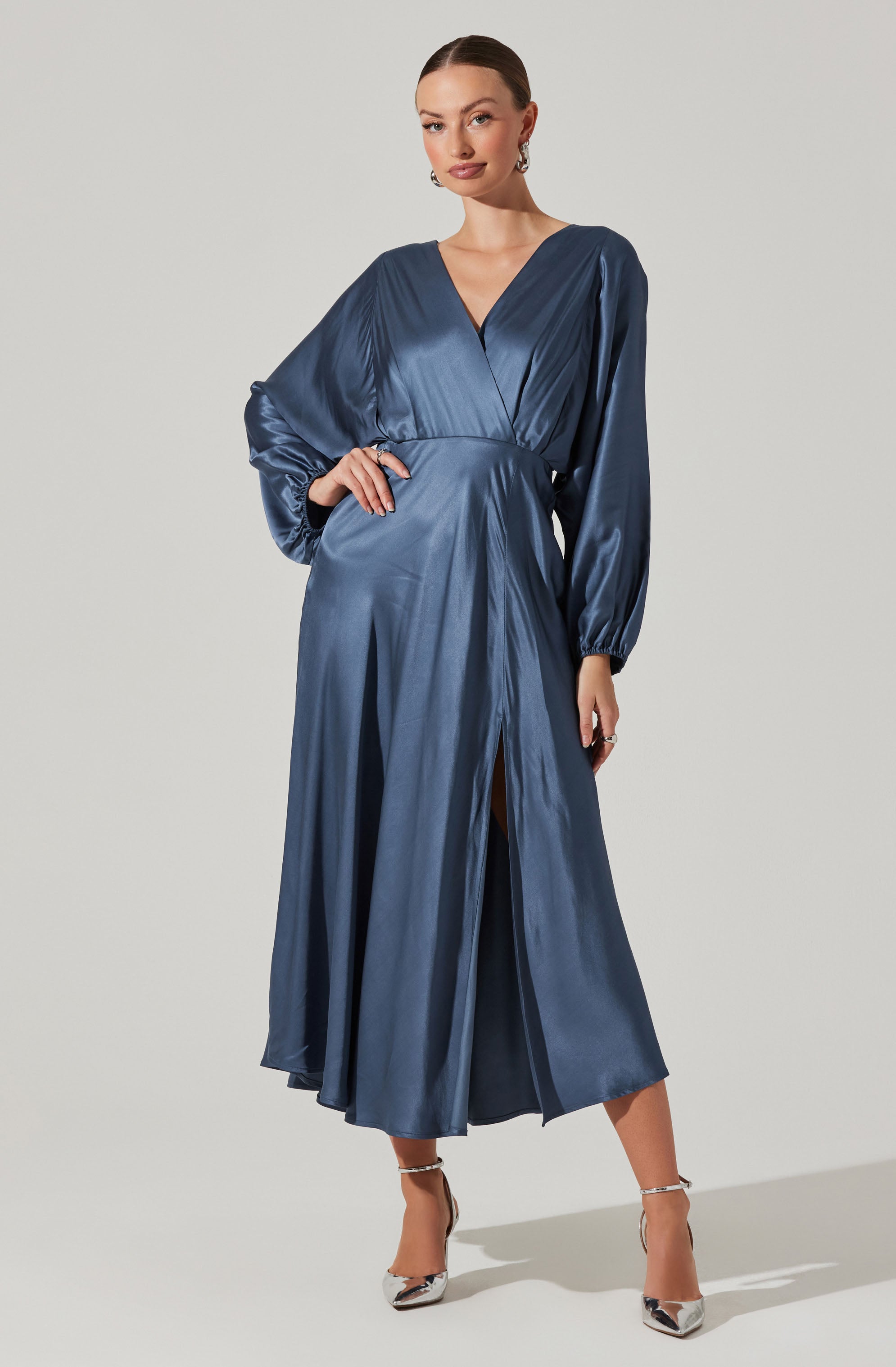 A woman is wearing a long, satin dress in a deep blue color. The dress has a V-neckline and long, loose sleeves gathered at the wrists. It features a high waistline and a front slit that reaches mid-calf, revealing a glimpse of her legs. She is accessorized with earrings and silver high-heeled shoes.