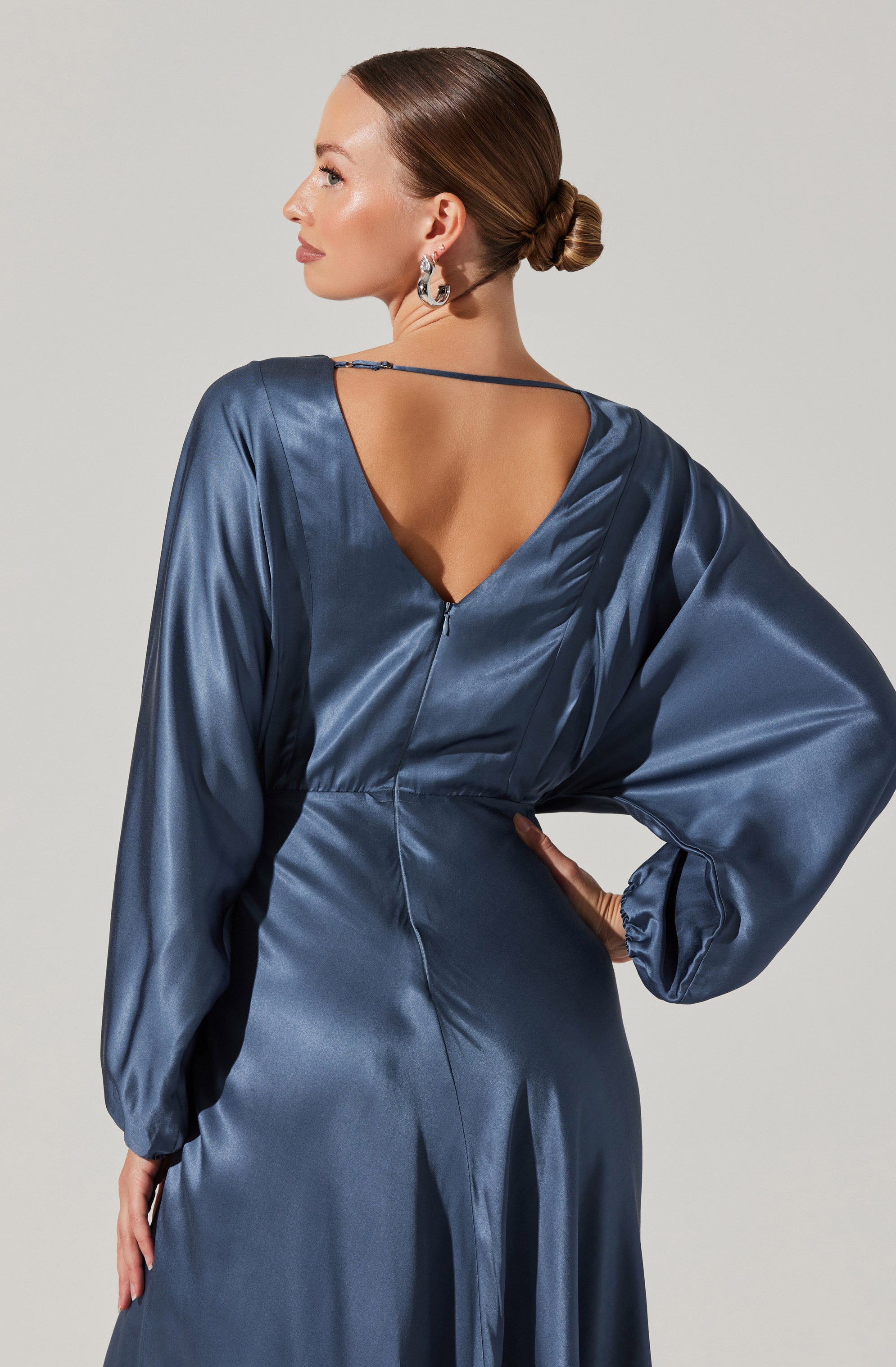 A woman is wearing a satin dress with long, puffed sleeves and a fitted waist. The dress features a deep V-shaped open back with a thin strap across the shoulders. The fabric has a shiny, smooth appearance. She has her hair styled in a low bun and is wearing large, patterned earrings.