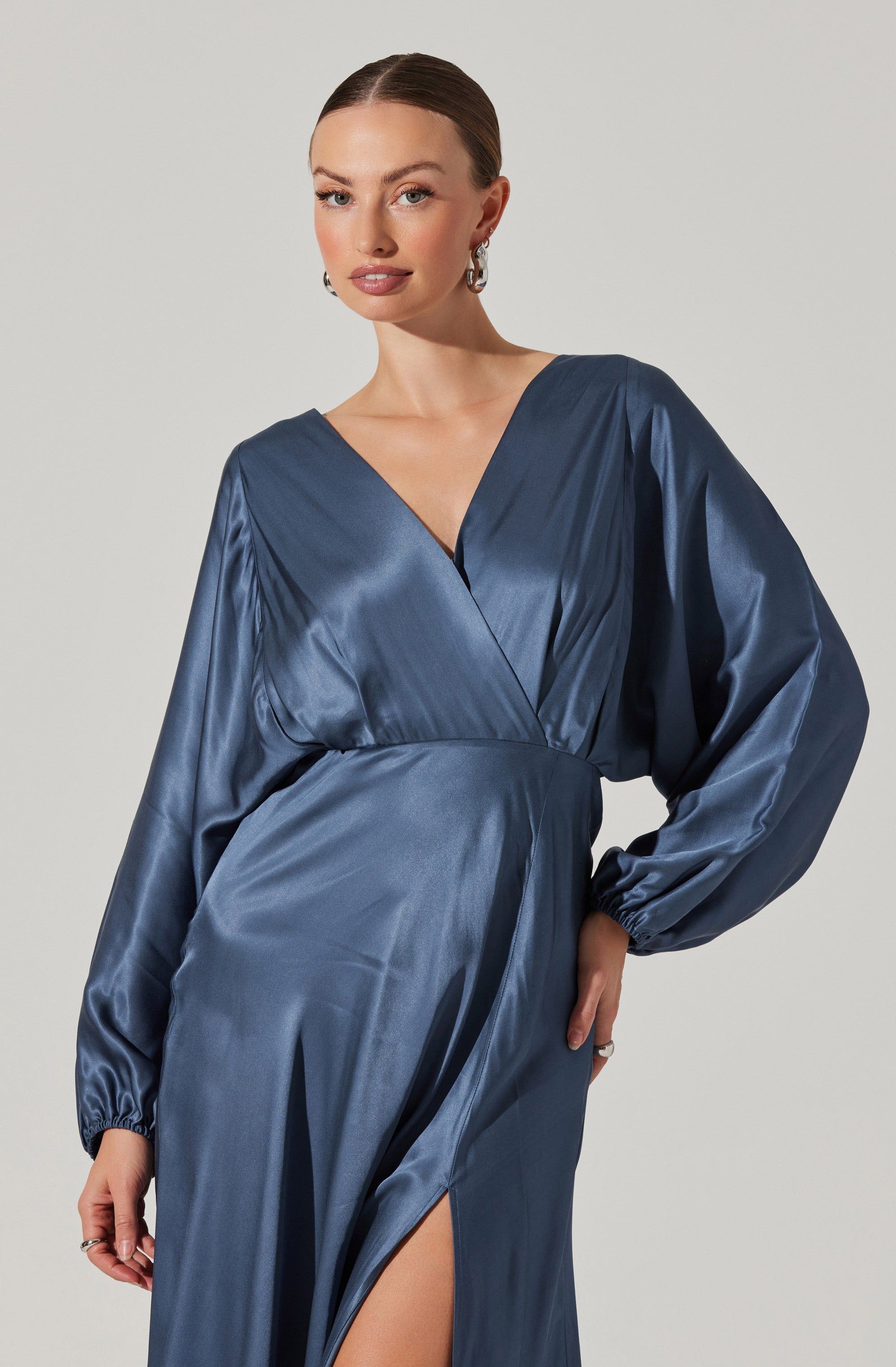 A woman is wearing a silky, satin-like long dress in a deep blue color. The dress features a V-neckline, long puffed sleeves with elastic cuffs, and a high slit on the side revealing part of her leg.
