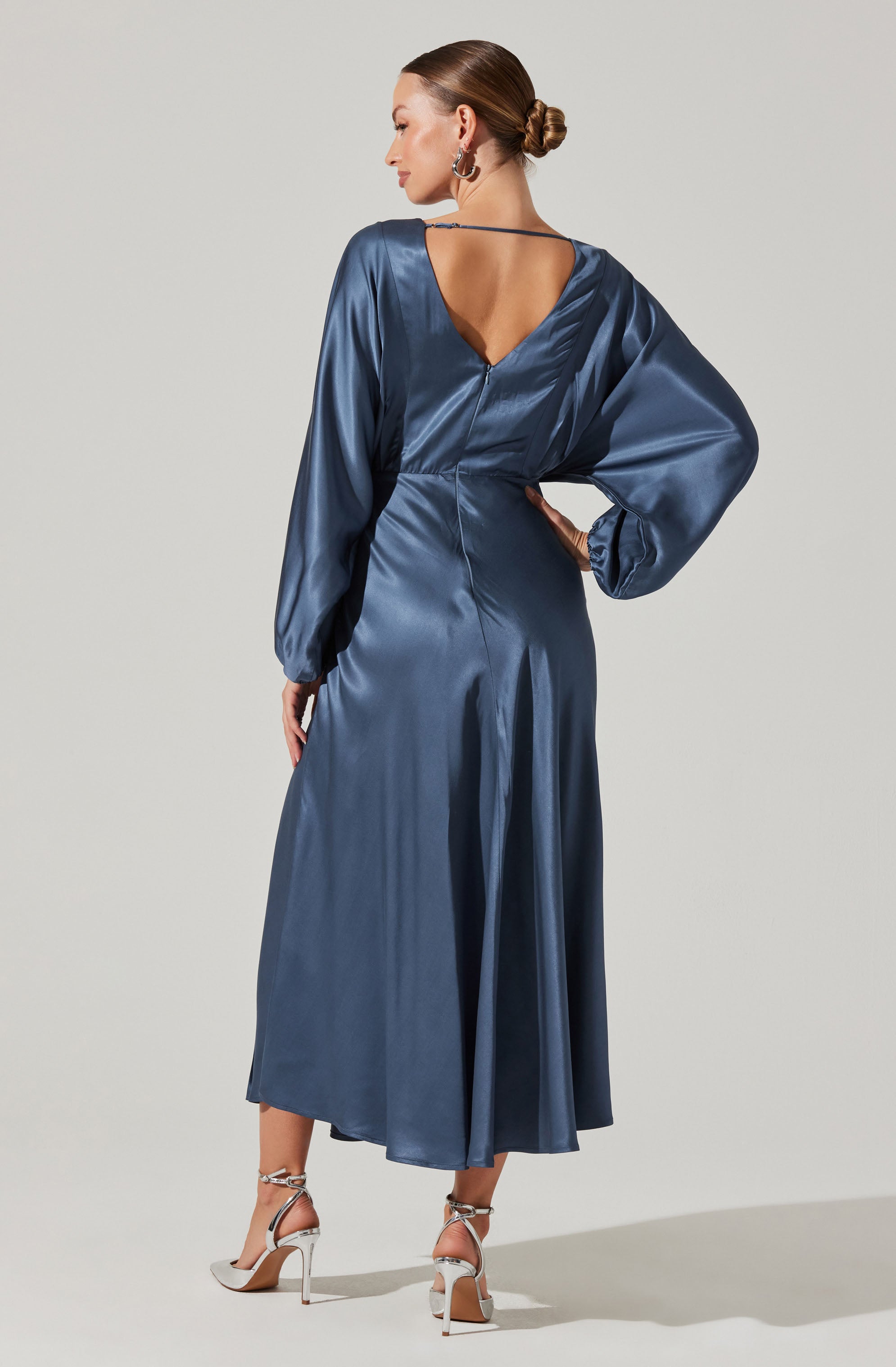 A woman is wearing a long, satin dress in a deep blue color. The dress features long, puffed sleeves with gathered cuffs, a V-shaped neckline at the back, and a fitted waist. She is also wearing high-heeled shoes with ankle straps.