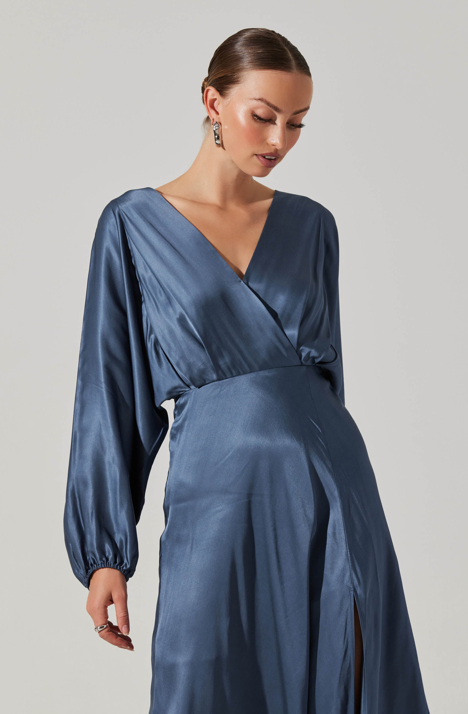 A woman is wearing a long, silky, navy blue gown with a V-neckline. The dress has a wrap-style bodice and long, slightly puffed sleeves gathered at the wrists. The skirt features a front slit.