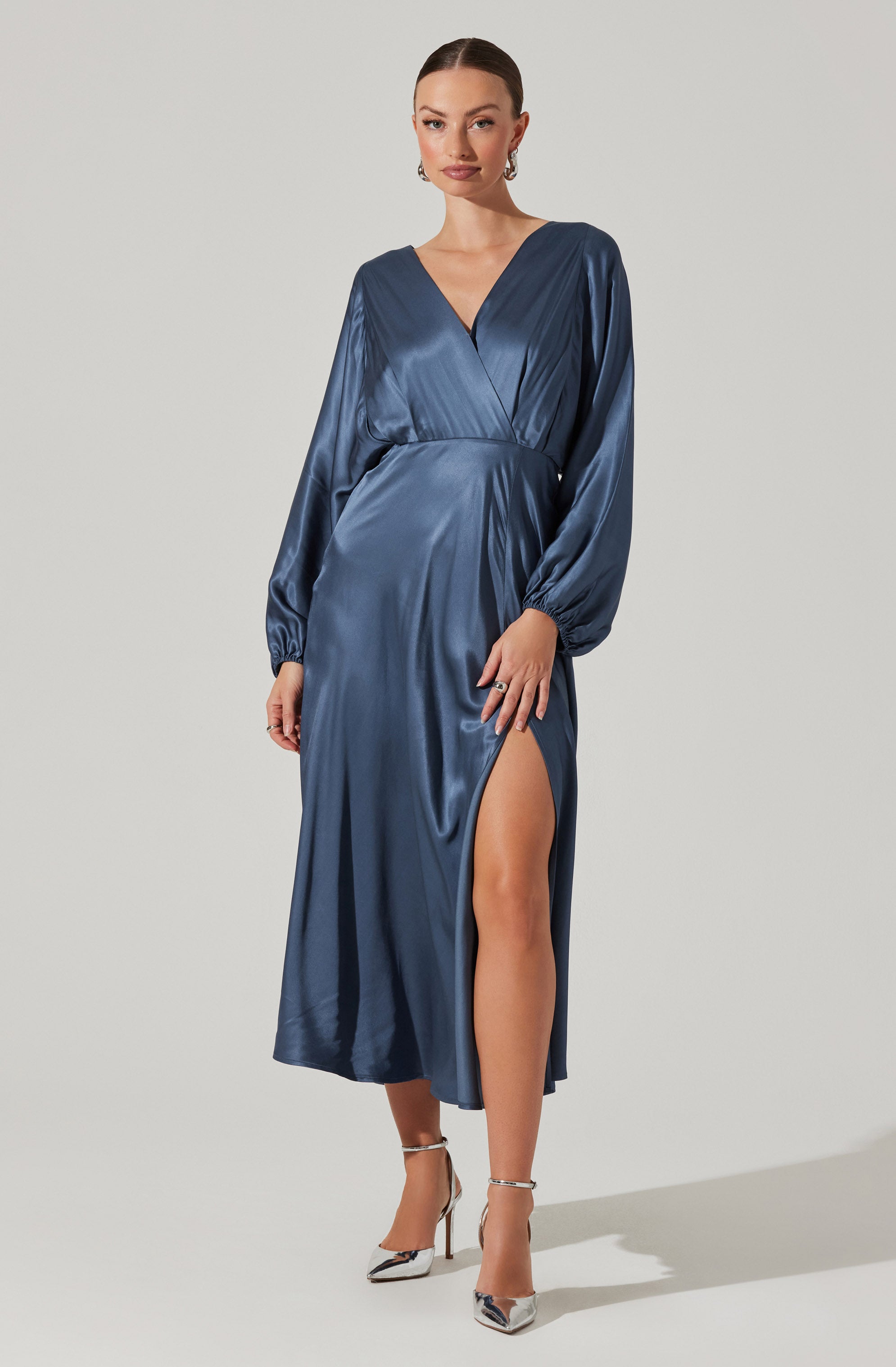 A woman is wearing a long, satin dress in a deep blue color. The dress features long, puffy sleeves gathered at the wrists, a V-neckline, and a high slit on the left side exposing her leg. She is also wearing metallic high-heeled shoes.