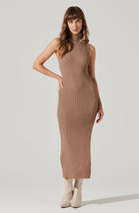 The woman is wearing a sleeveless, high-neck, ribbed knit dress in a tan or beige color. The dress is form-fitting and extends to her ankles. She is also wearing light-colored, pointed-toe ankle boots.
