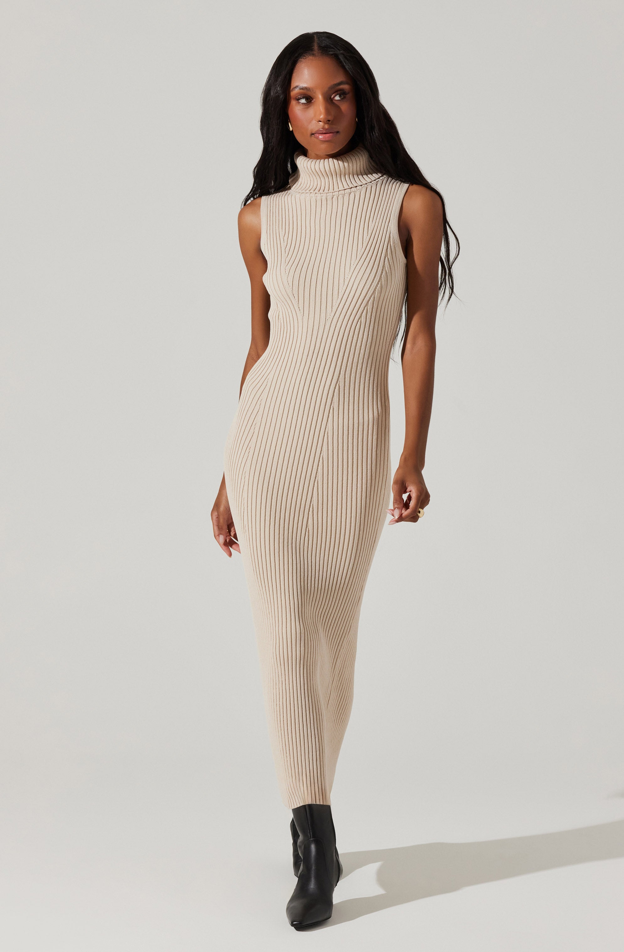 A woman is wearing a sleeveless, high-neck, beige ribbed knit dress that extends to her ankles. She has on black ankle boots and is standing against a plain, light gray background.
