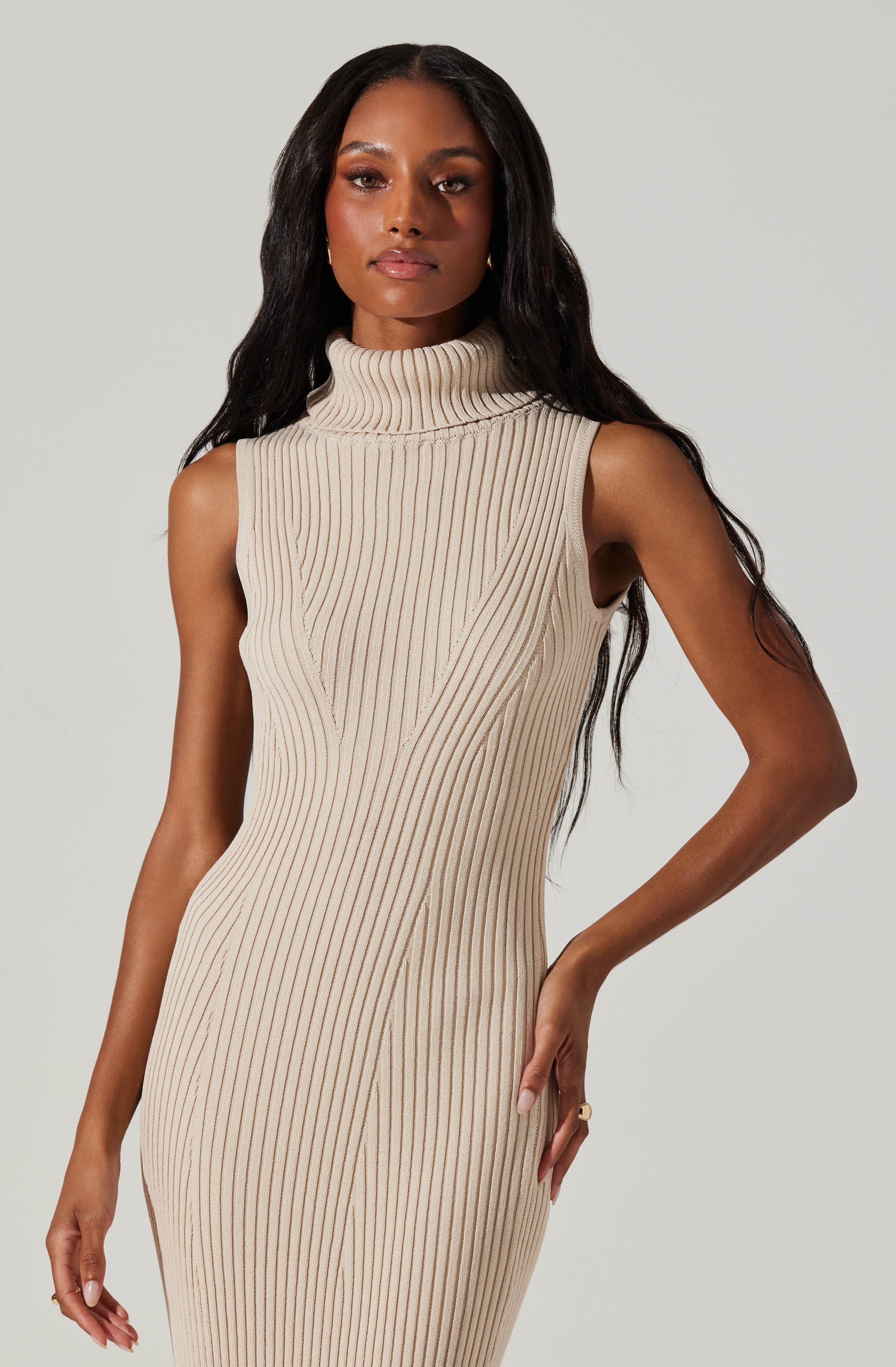 A woman is wearing a sleeveless, beige ribbed knit dress with a high turtleneck collar. The dress features vertical ribbing and has a fitted silhouette.