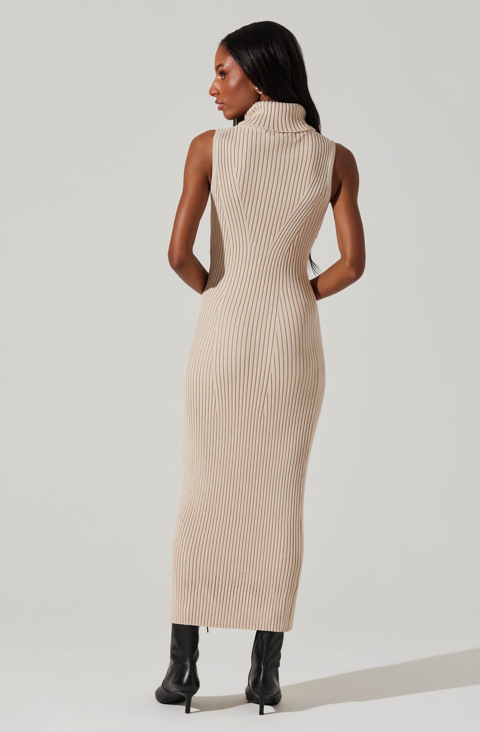 A woman is wearing a sleeveless, beige, ribbed knit dress that is form-fitting and ankle-length. The dress features a turtleneck collar and has vertical, slightly wavy ribbing throughout. She is also wearing black high-heeled boots.